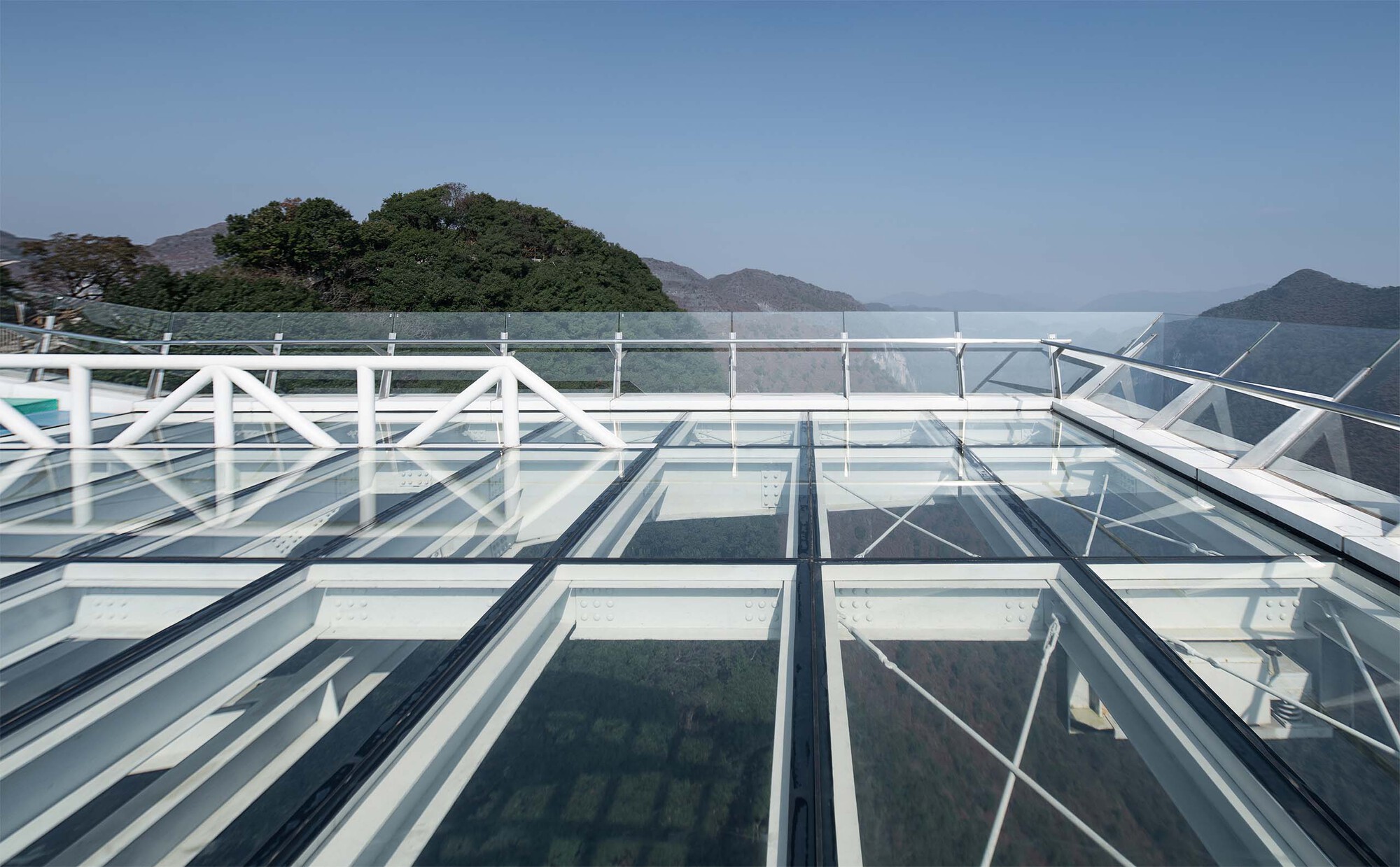 Glass floor panels supported by white steel trusses with mountain ridges visible beyond the railing