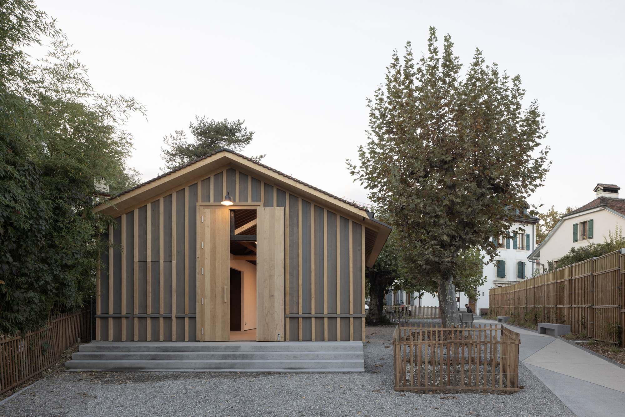Gabled volume with vertical timber and panel cladding facing a gravel courtyard with a central tree