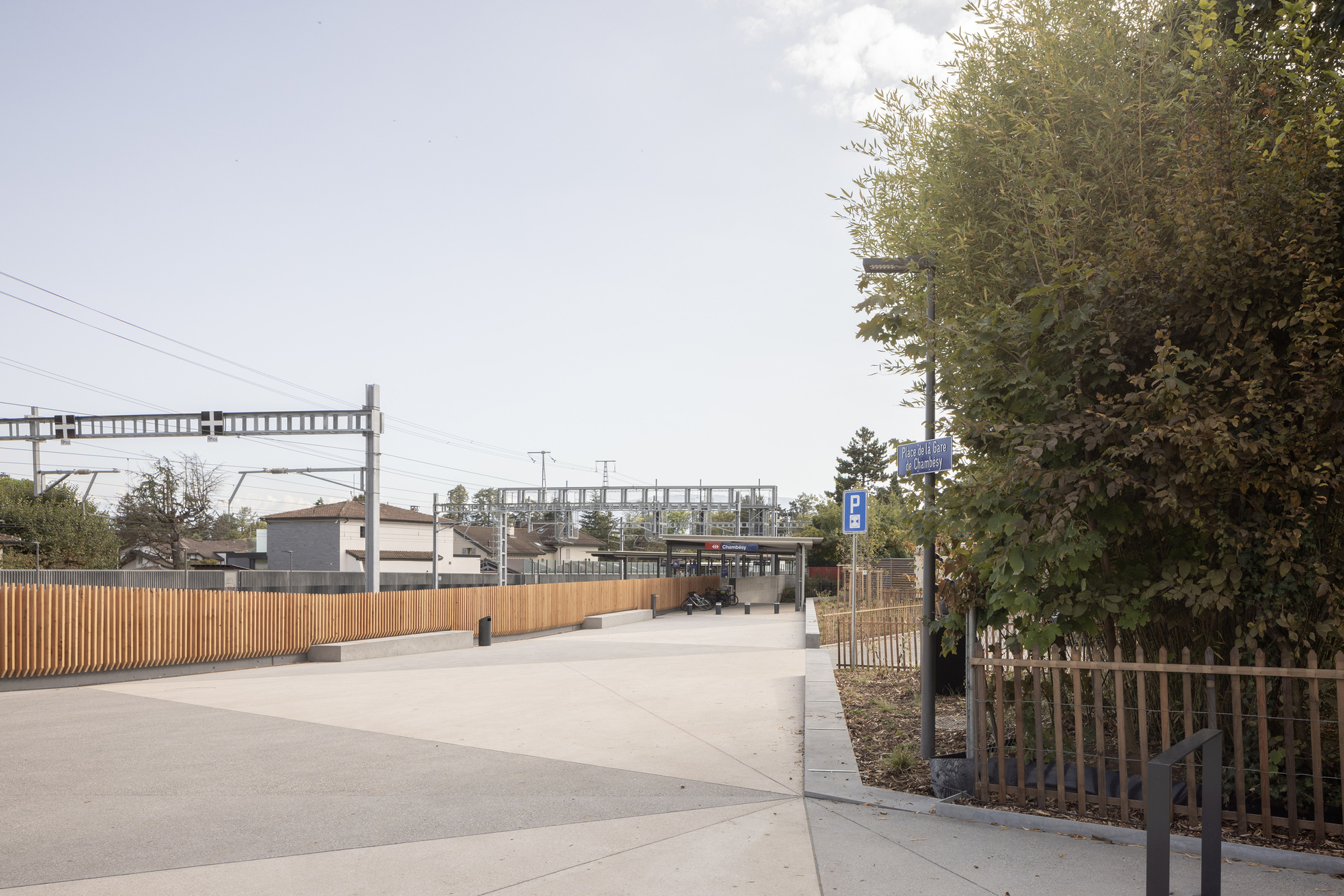 Public plaza with timber-screened walkway leading toward a railway footbridge in overcast daylight