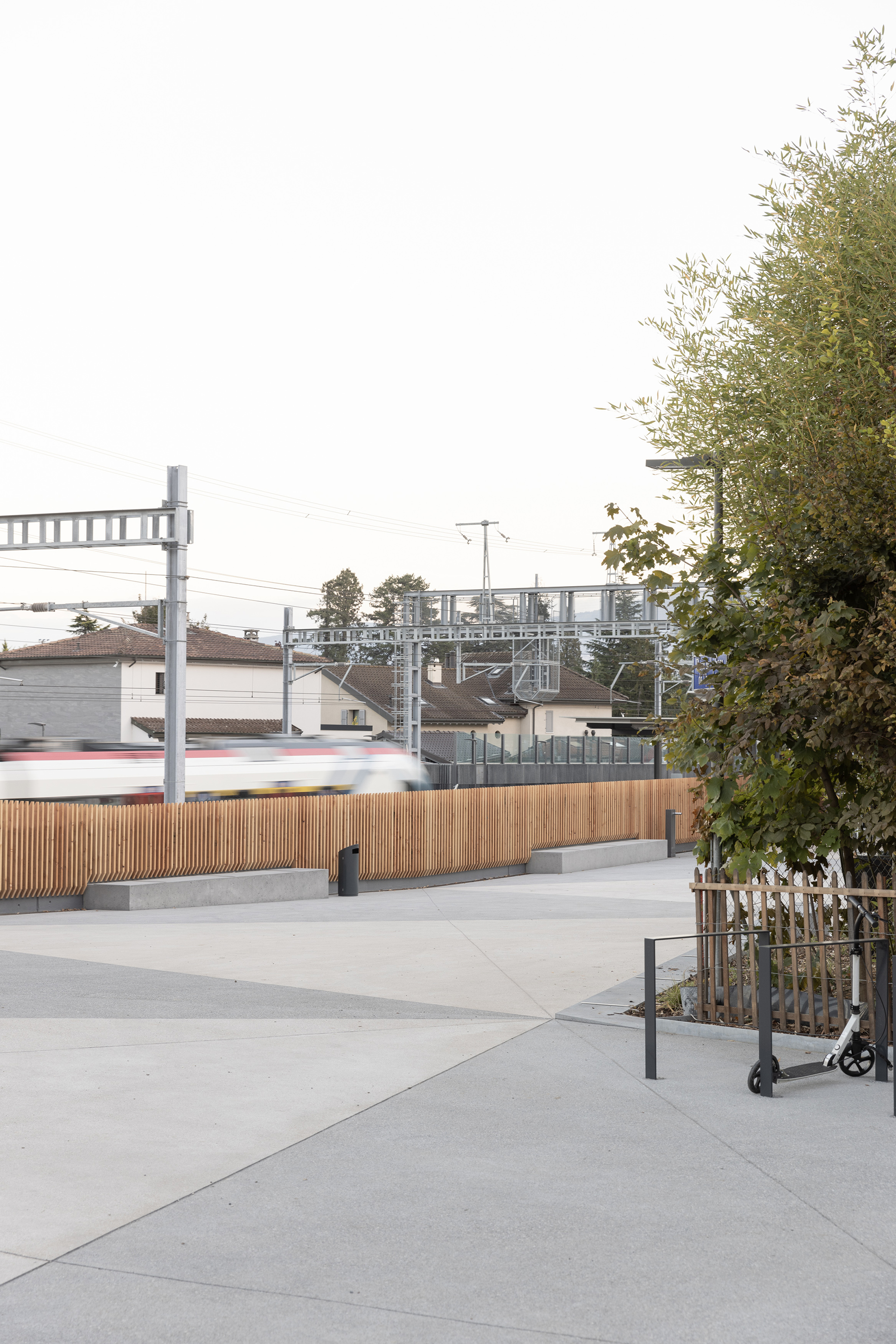 Concrete plaza with timber screen wall beside passing train and scooter parked near corner