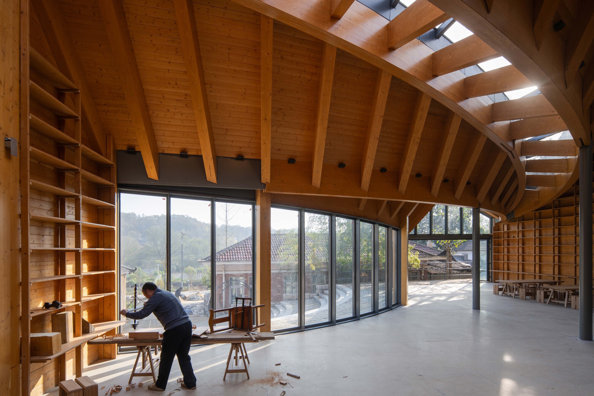 Interior space with exposed timber beams and skylights, a person working at a table near floor-to-ceiling glazing
