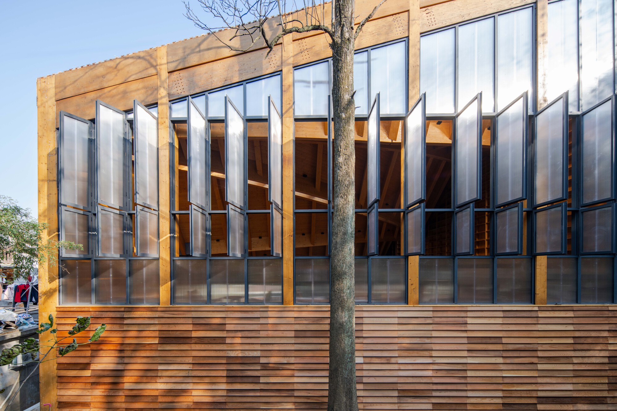 Exterior facade combining horizontal wood slats with pivoting polycarbonate shutters beside a bare tree trunk