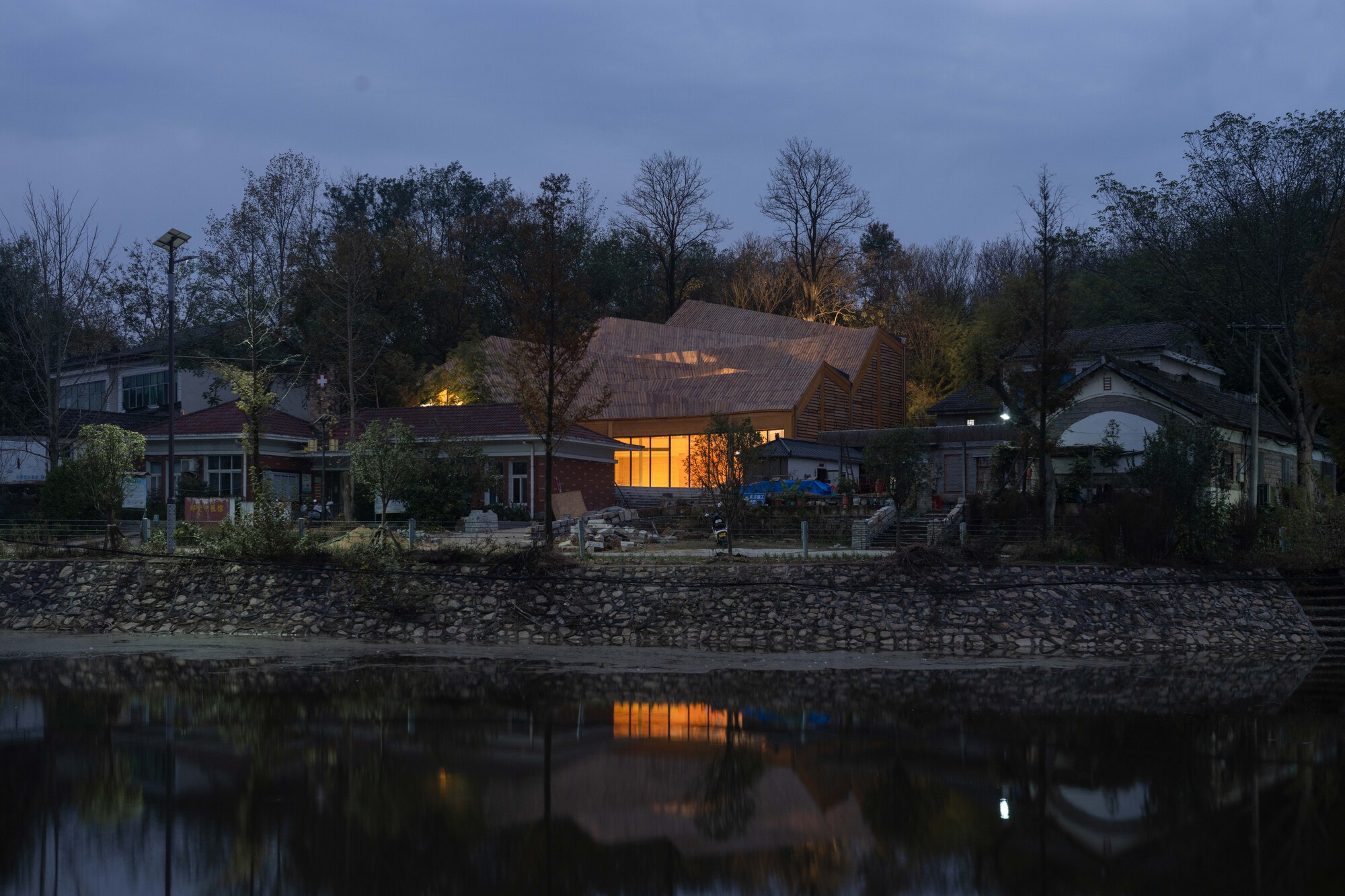 Waterfront view of the shingled roof and illuminated interior at dusk