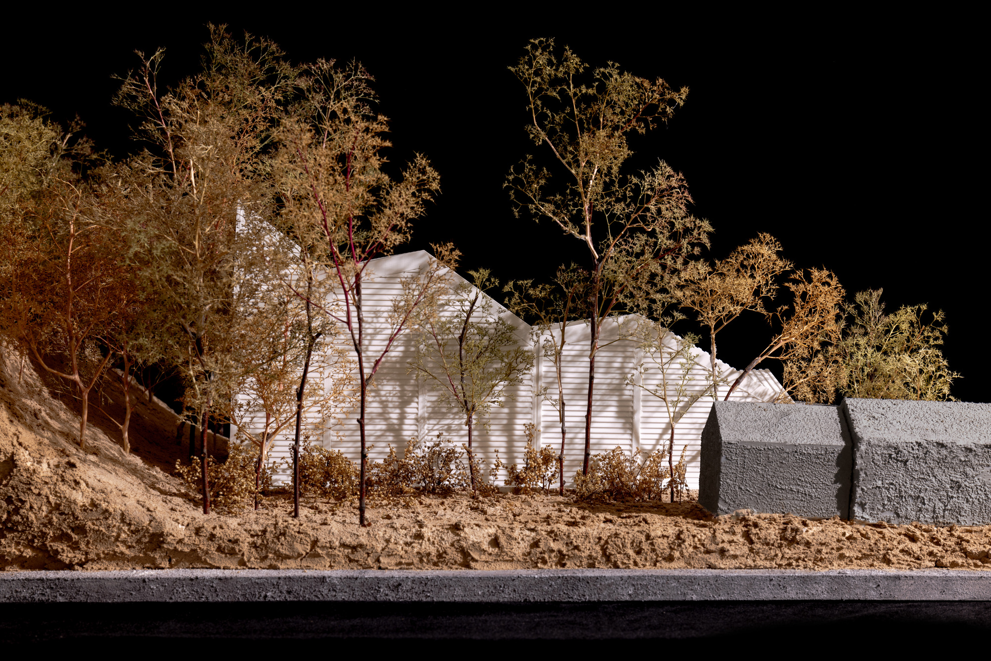 Side view of physical model showing illuminated ribbed facade behind bare trees and textured retaining wall