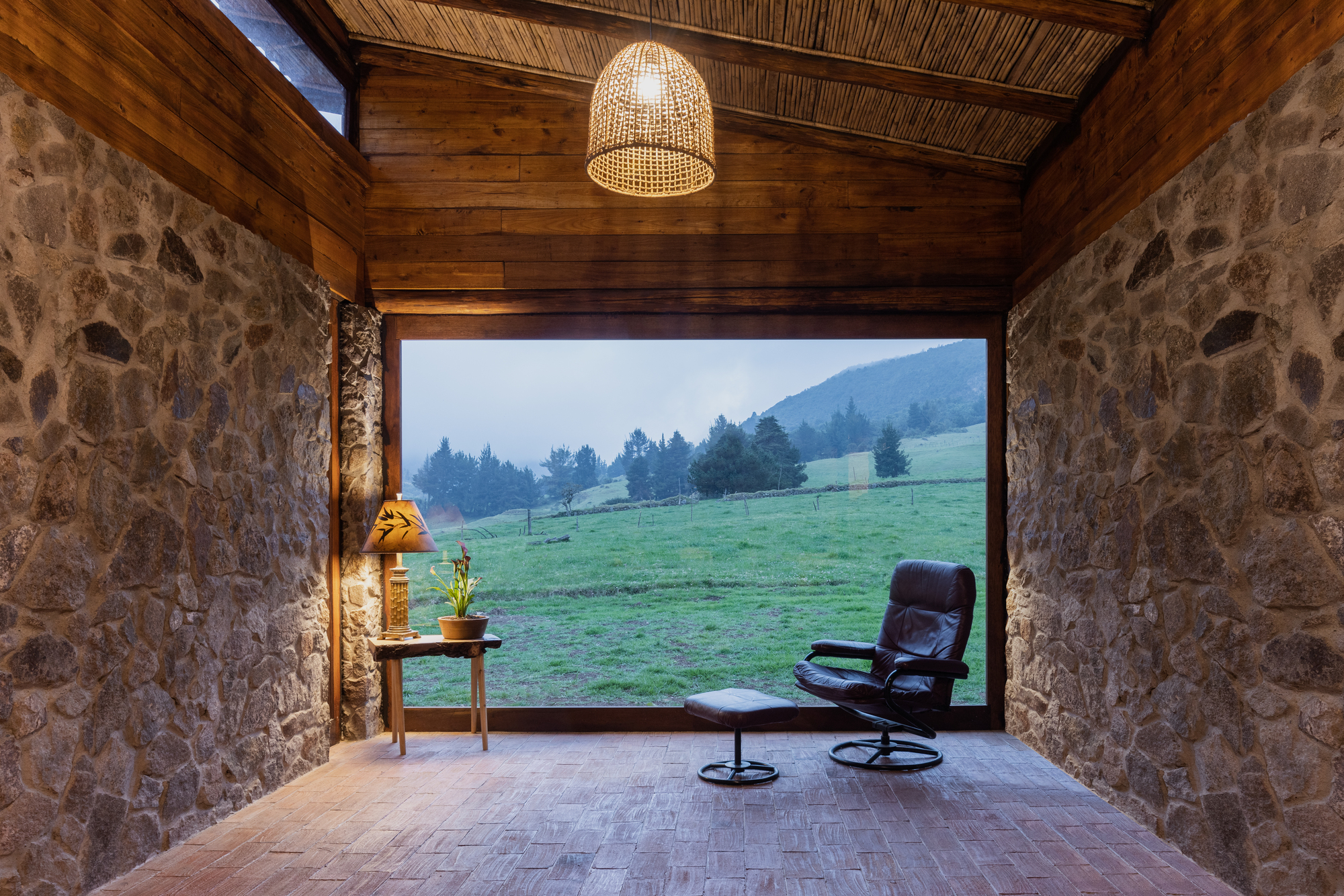 Stone walled room with woven bamboo ceiling and wicker pendant light framing view to misty pasture at dusk