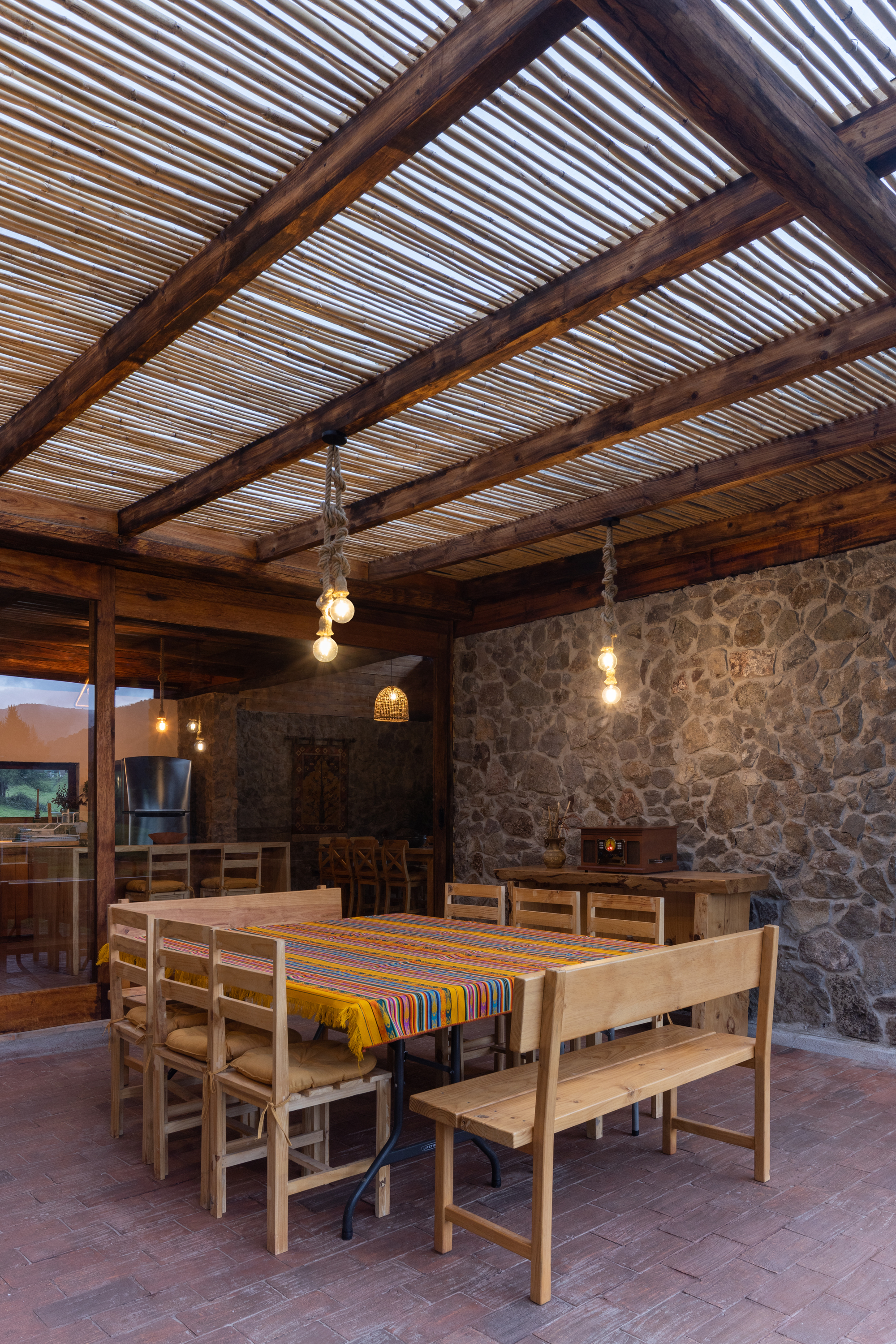 Outdoor dining area with slatted timber ceiling, stone masonry walls and pendant bulbs at dusk