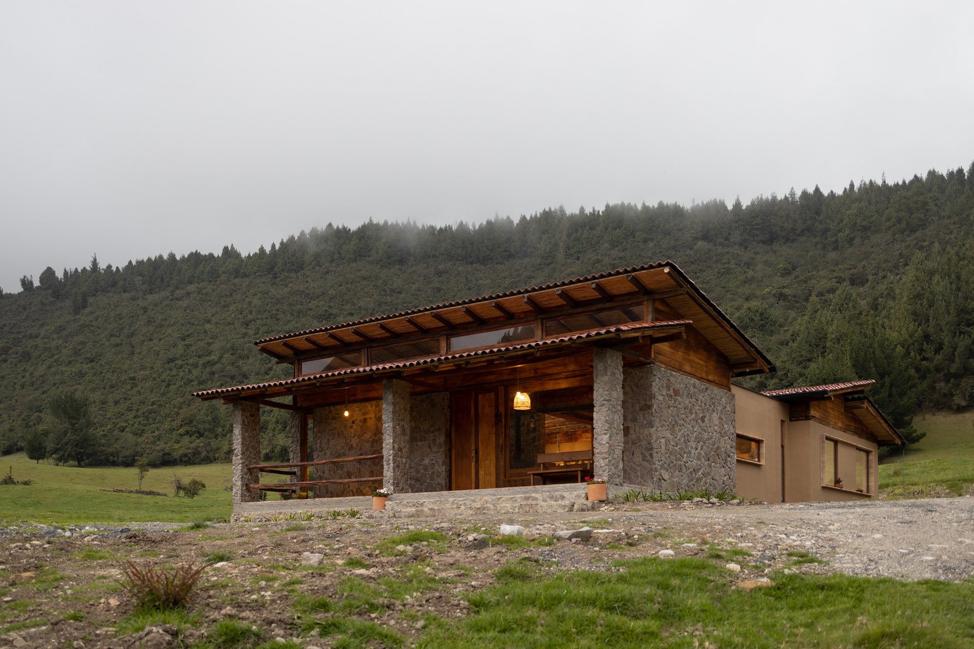 Low-slung residence with stone columns and timber frame set in meadow below forested hillside in mist