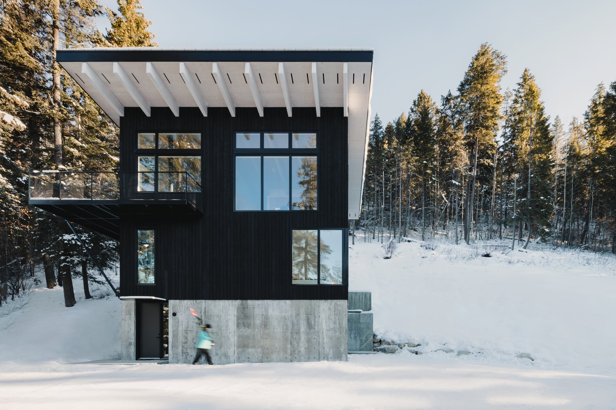 Black timber clad volume with gridded windows on a concrete base in a snow covered forest with a figure passing by