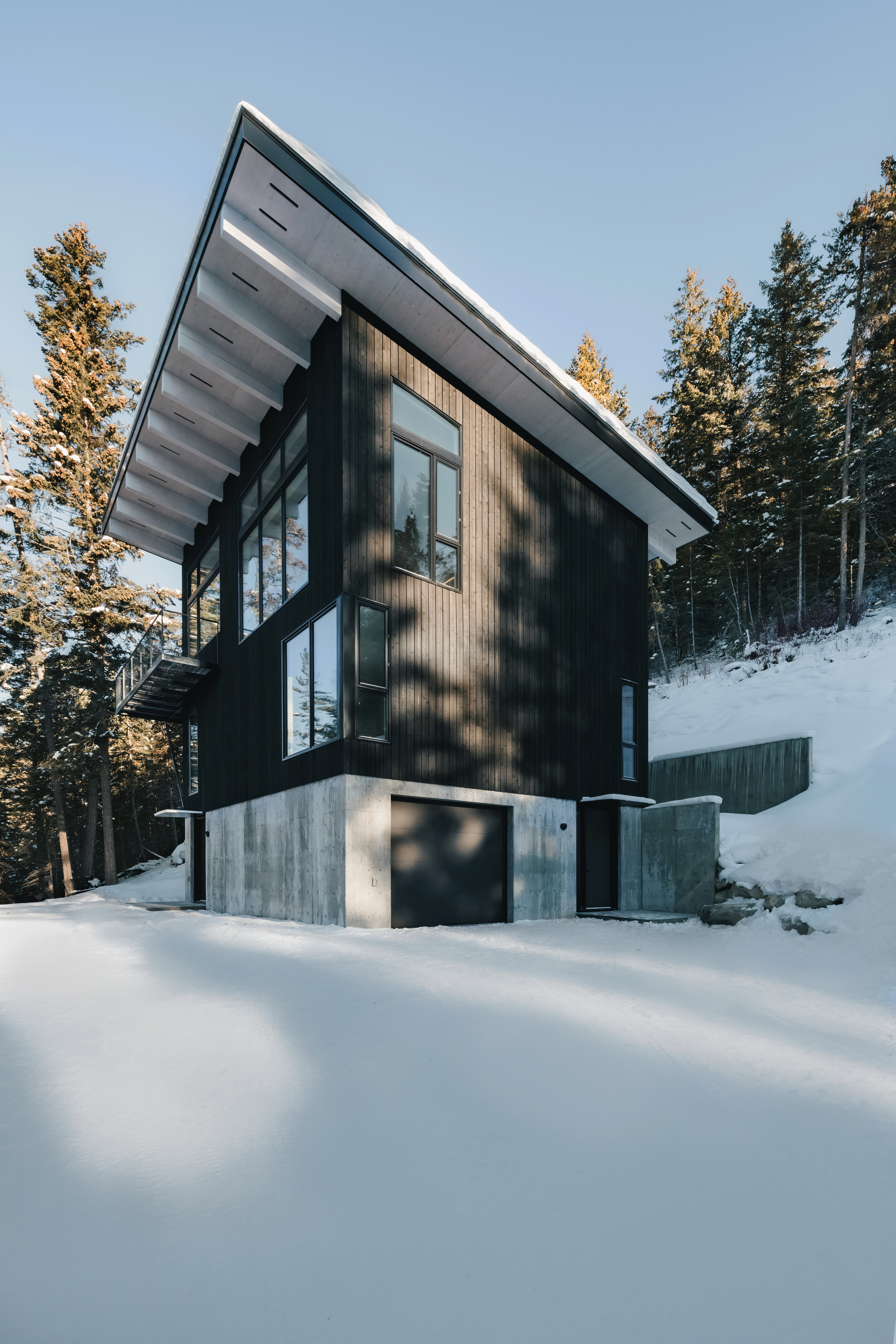 Two-storey facade showing dark cladding on exposed concrete plinth surrounded by deep snow and pine trees in afternoon light