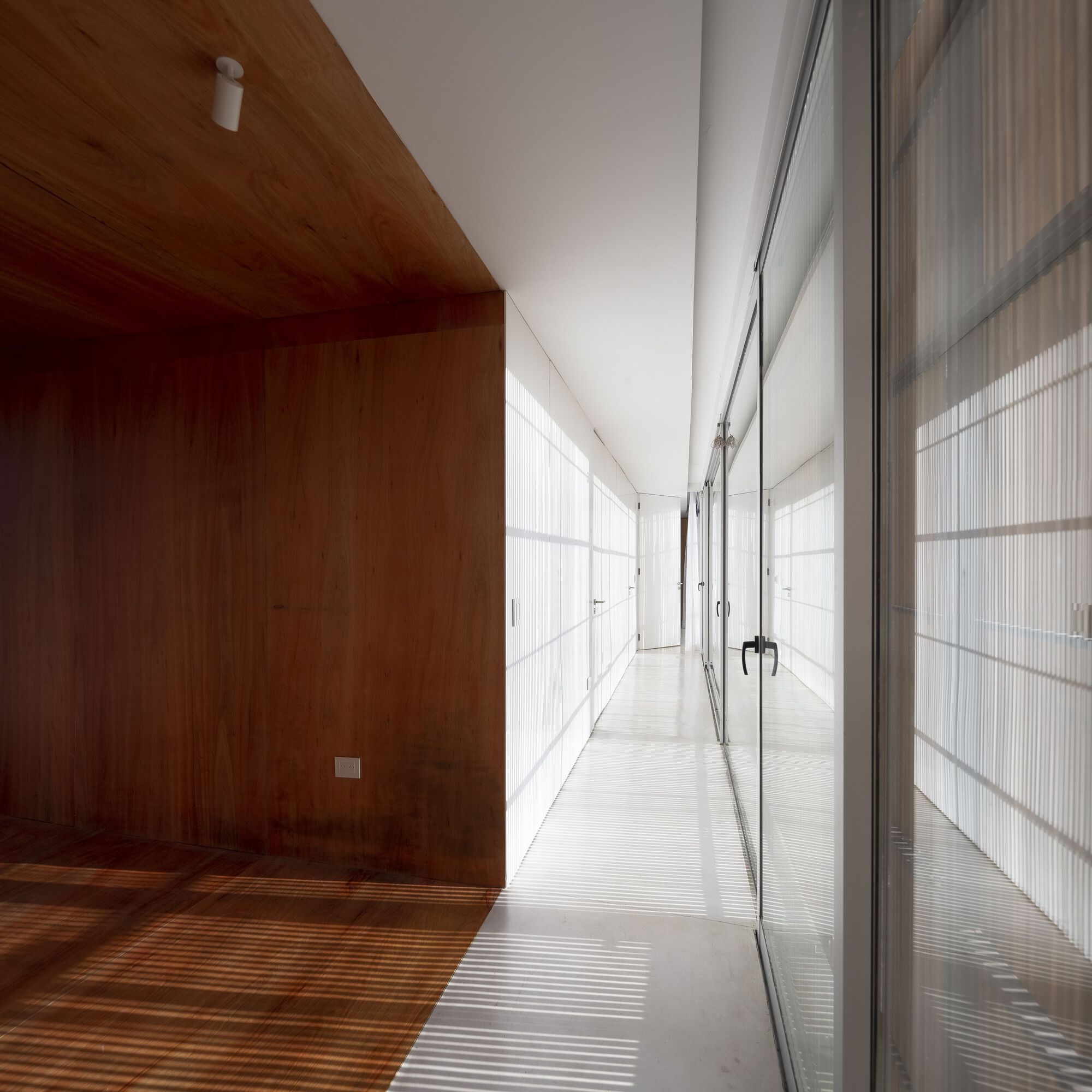 Interior corridor with timber-clad walls and ceiling alongside translucent sliding screens casting linear shadows