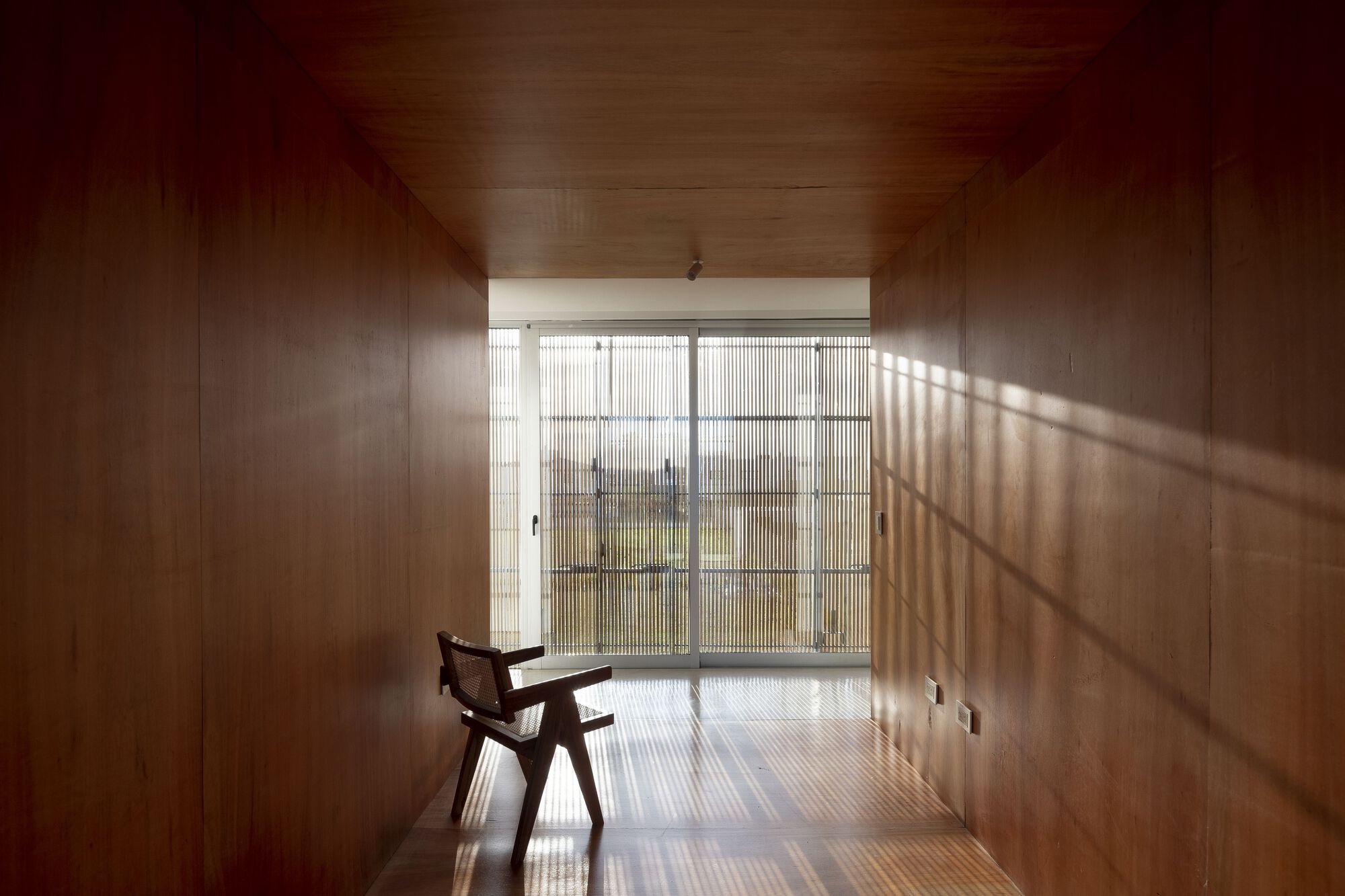 Wood-paneled room with a single chair facing a translucent glazed wall filtering diagonal sunlight
