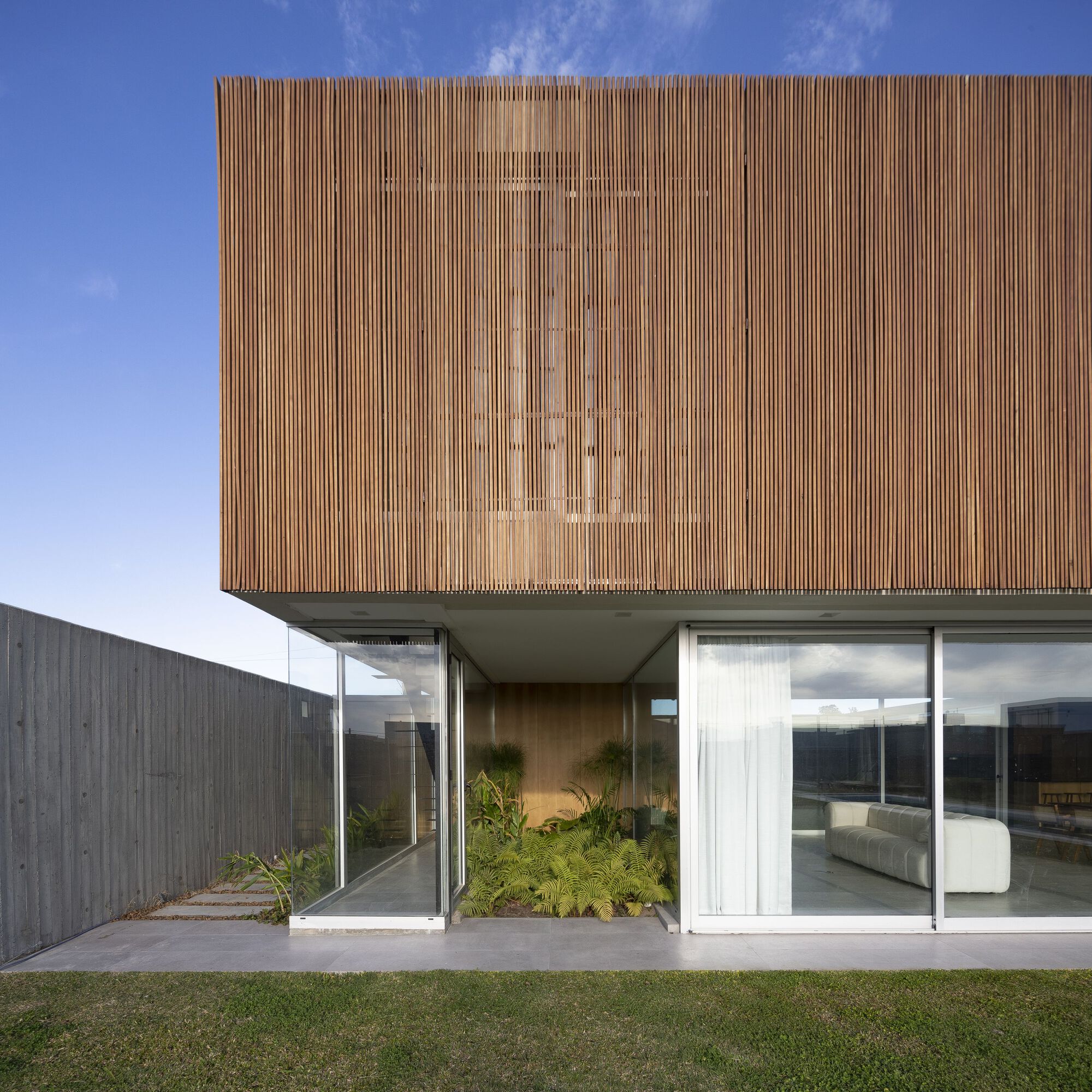 Cantilevered timber-clad volume above glass-walled ground floor with courtyard planting and lawn