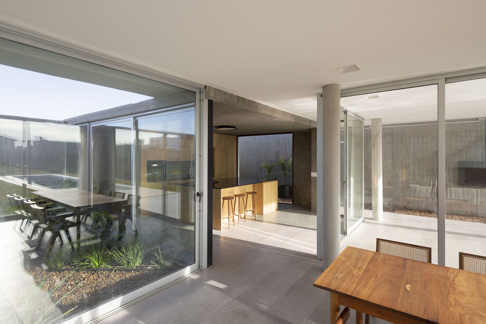 Glass-enclosed courtyard with planted bed adjoining an interior kitchen and timber dining table
