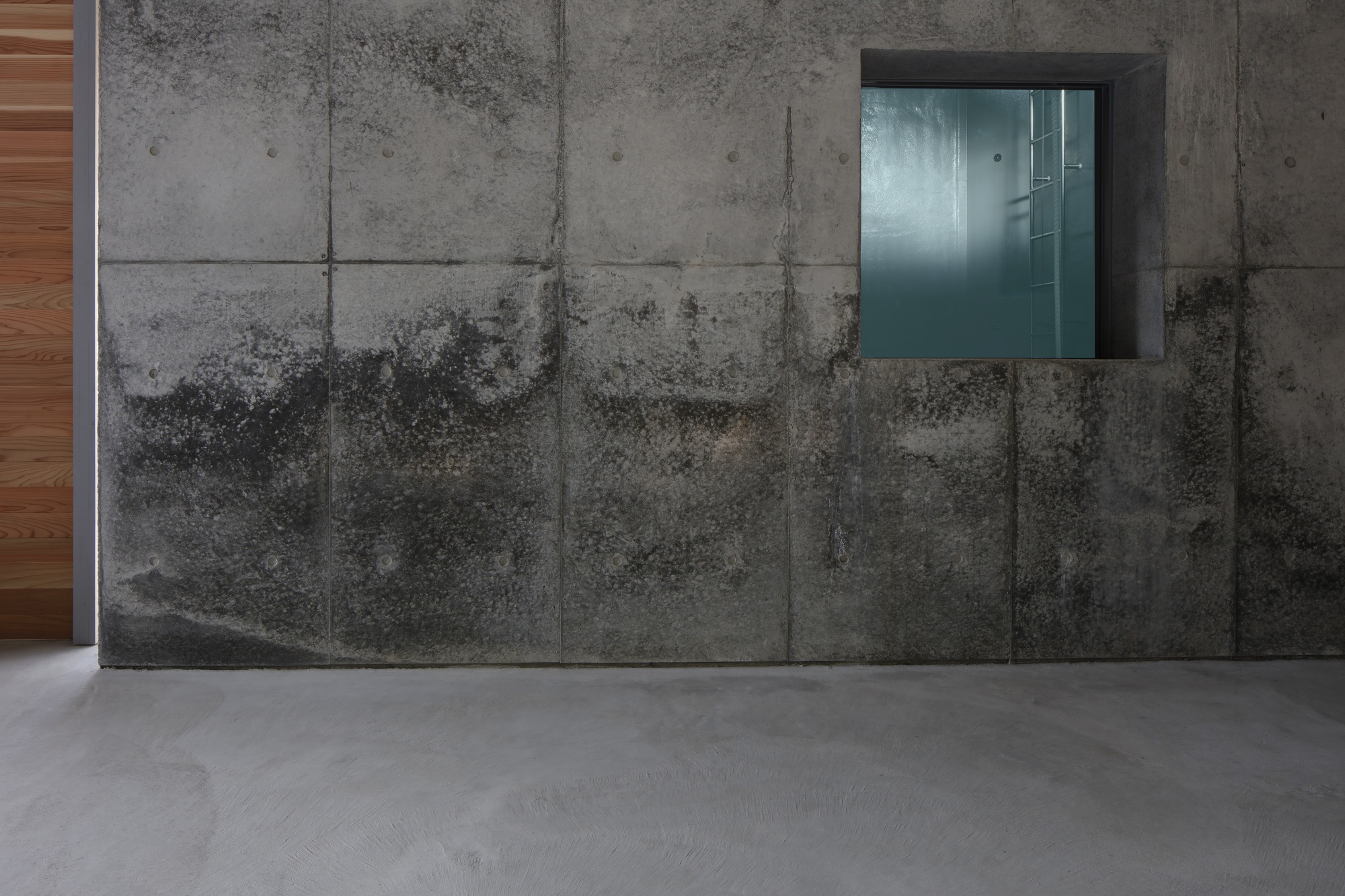Weathered concrete panel wall with recessed stainless steel window opening and polished grey floor