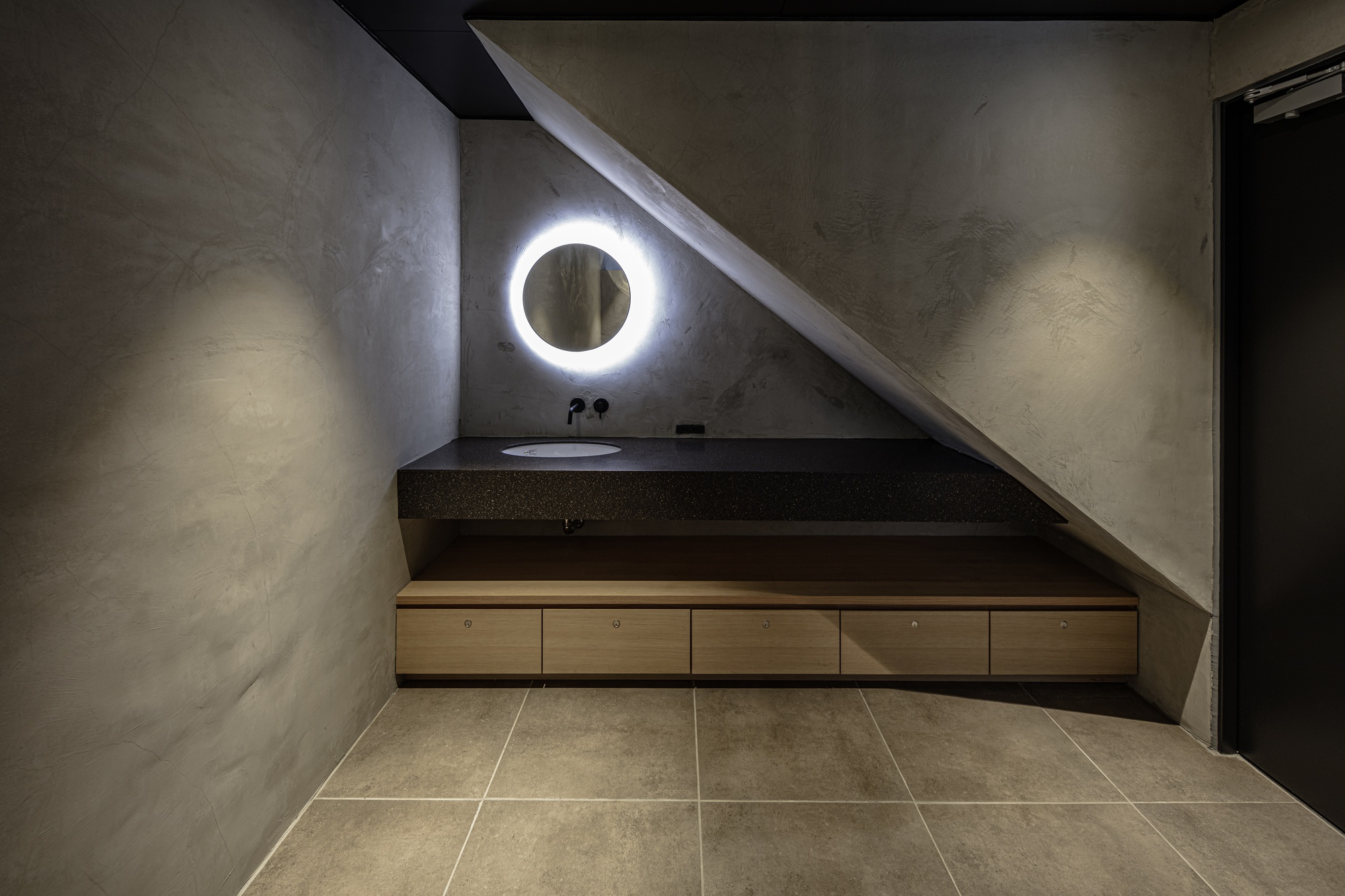 Bathroom vanity tucked beneath a sloped ceiling with circular illuminated mirror at night