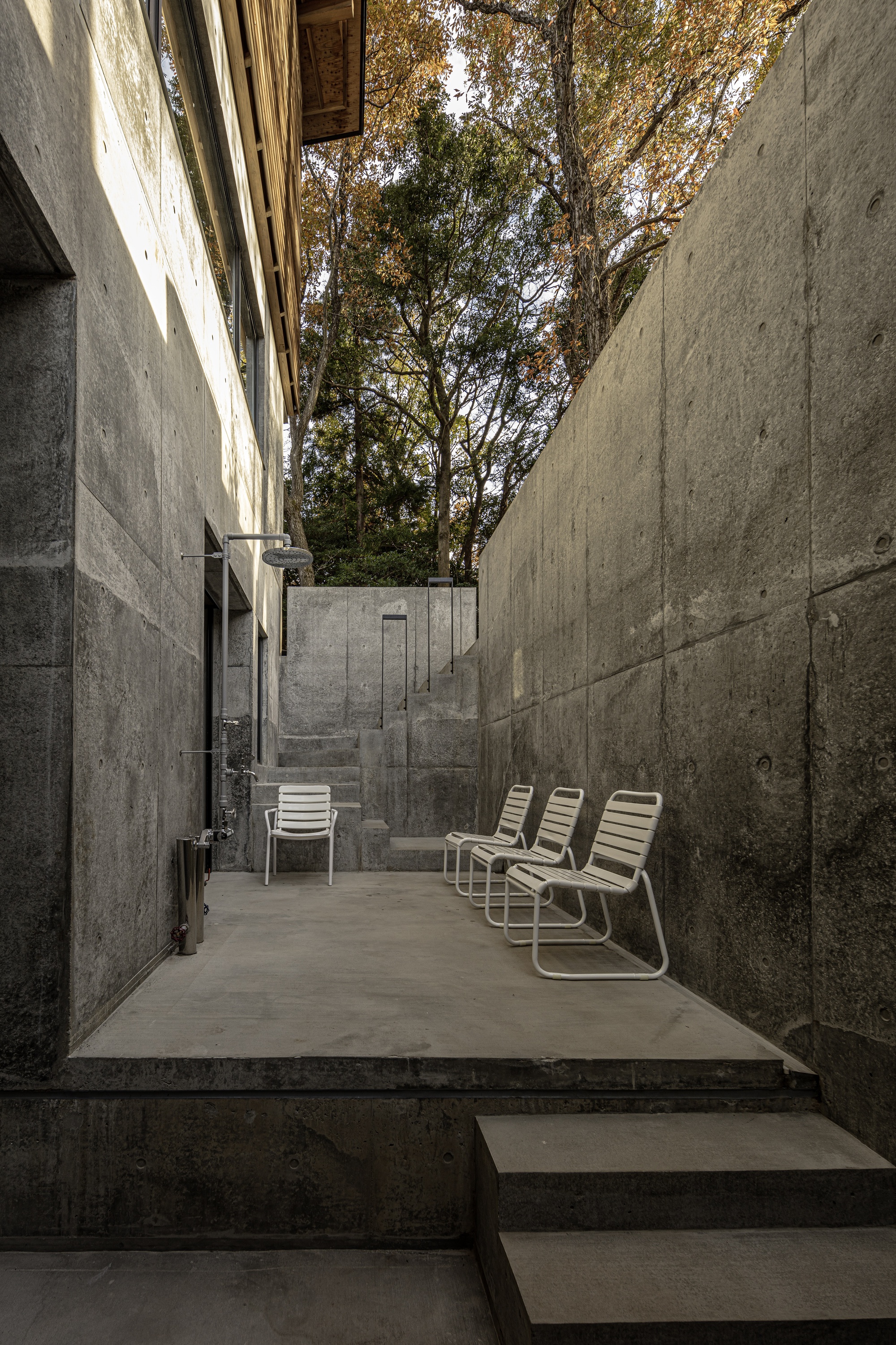 Concrete courtyard with metal lounge chairs flanked by board-formed walls beneath autumn trees