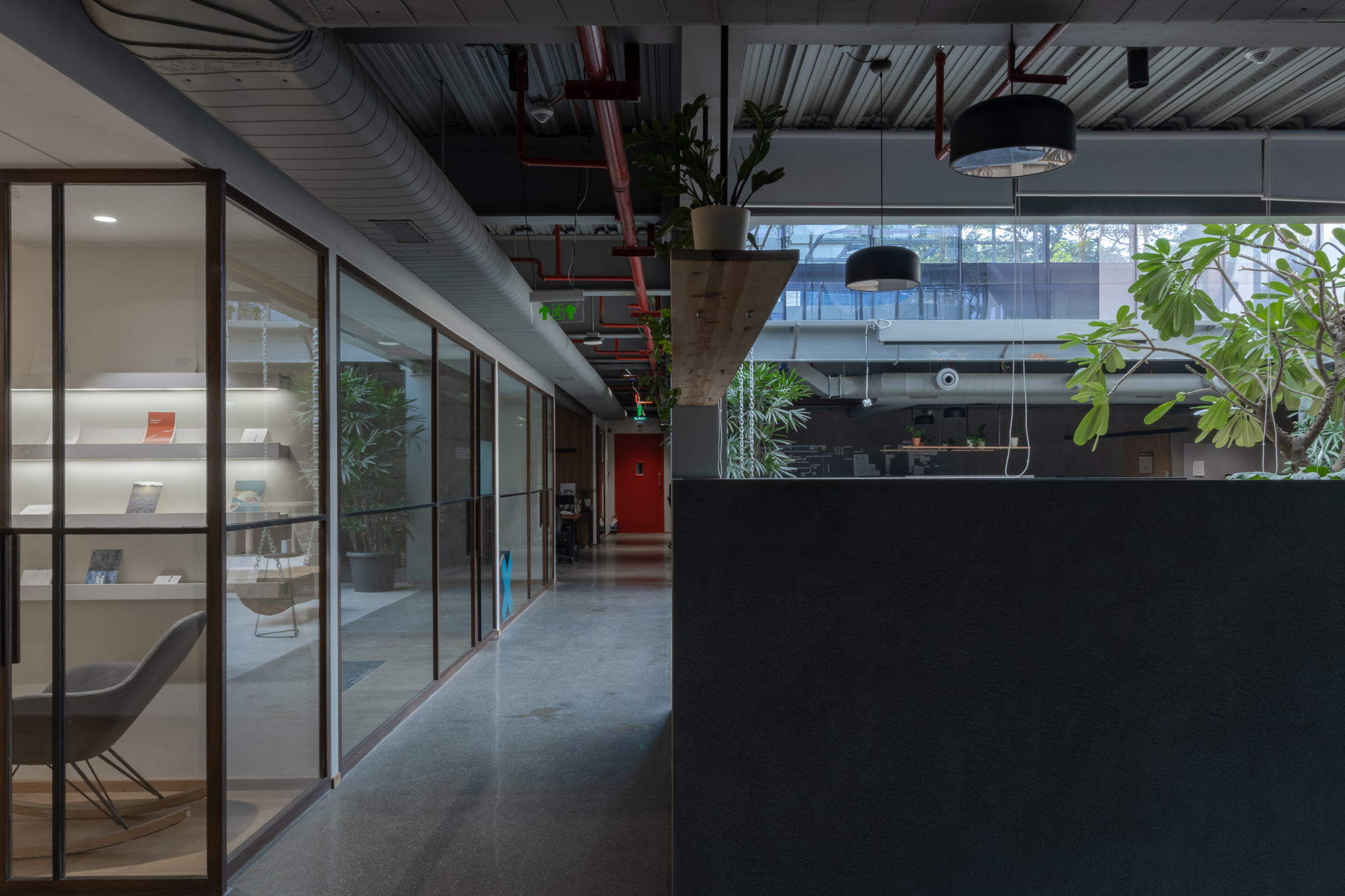 Office corridor with exposed ceiling ducts, glazed meeting rooms, and potted plants along the passage