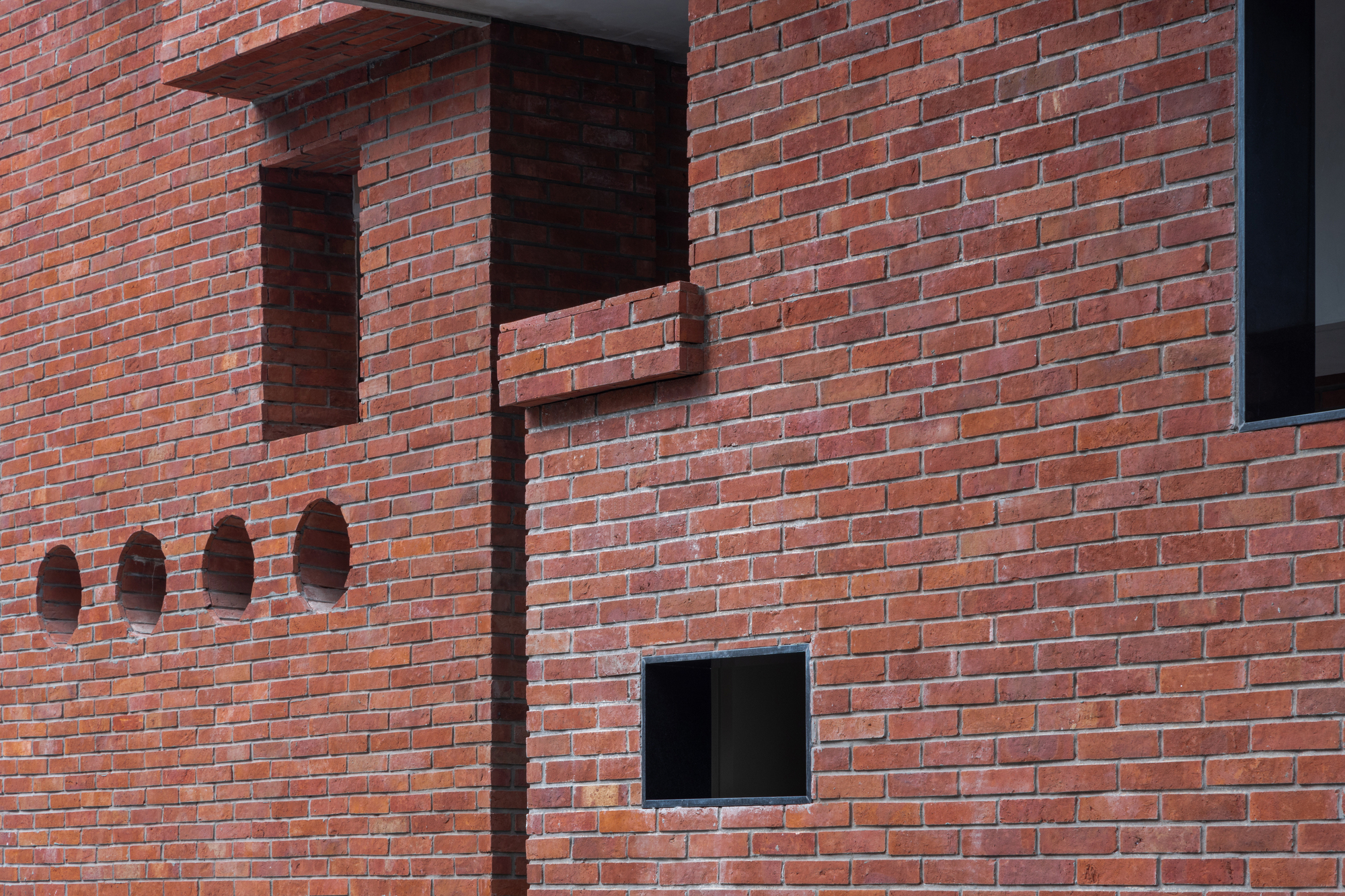 Red brick wall with projecting corbelled elements, recessed niches, and square window openings in afternoon light