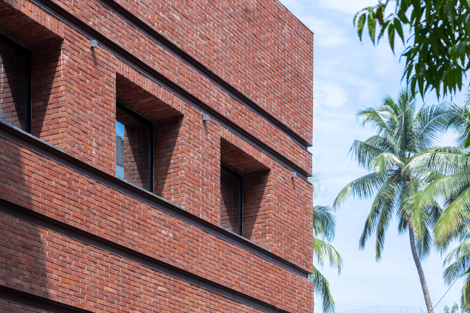 Stacked brick facade with tapering arched openings and horizontal banding against palm trees and blue sky
