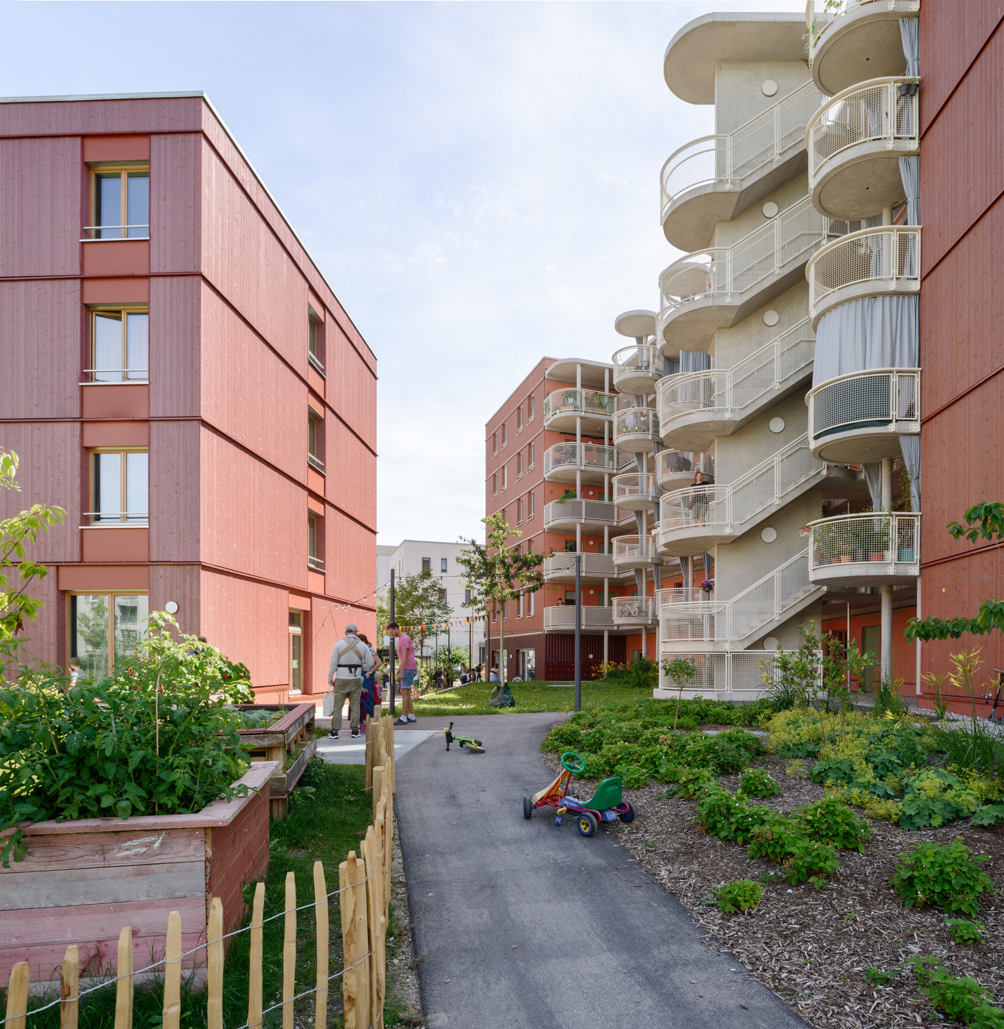 Residential courtyard path between planted gardens with a tricycle and residents gathering in late afternoon light