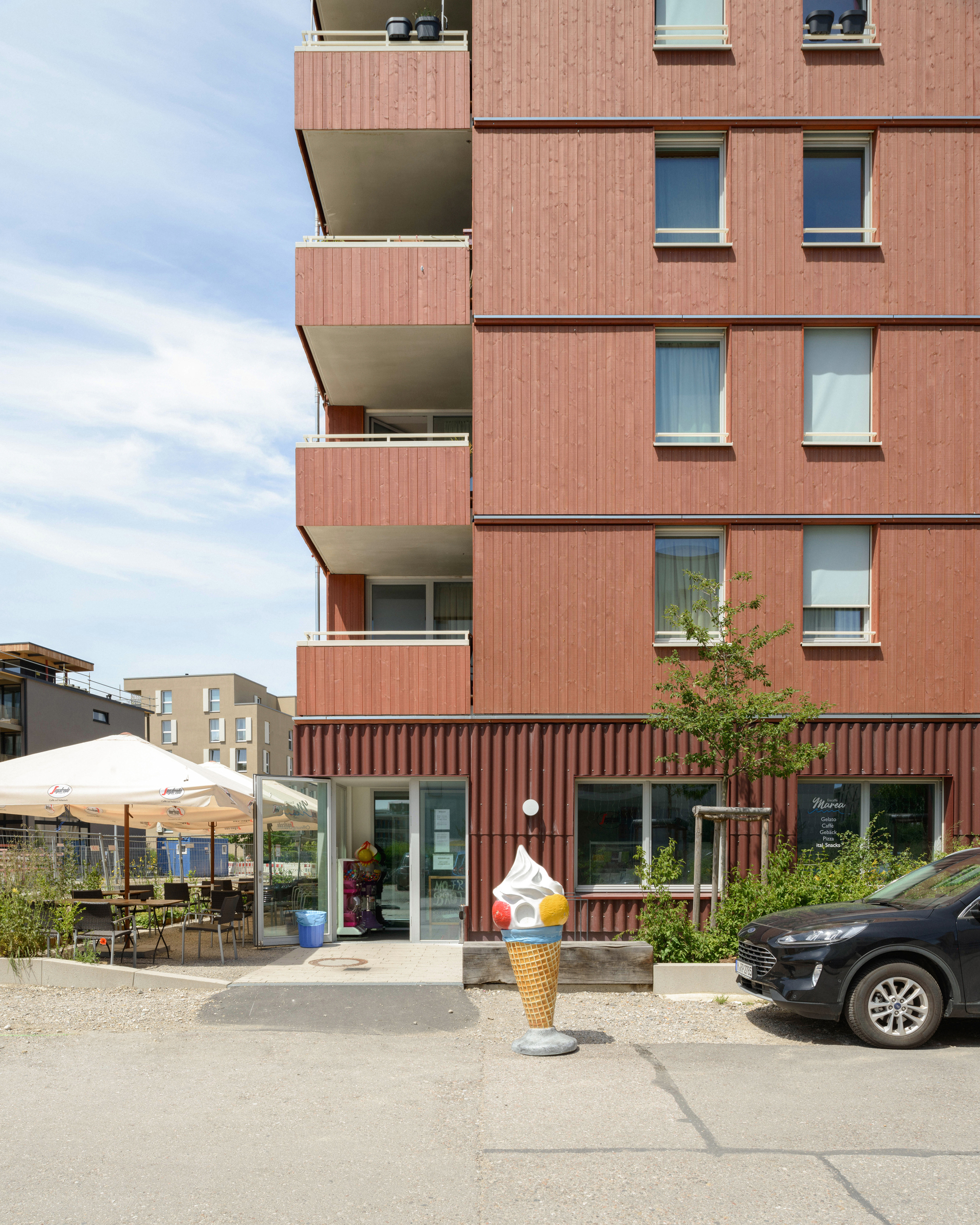 Ground floor entrance with corrugated cladding, outdoor cafe seating, and oversized ice cream cone sculpture