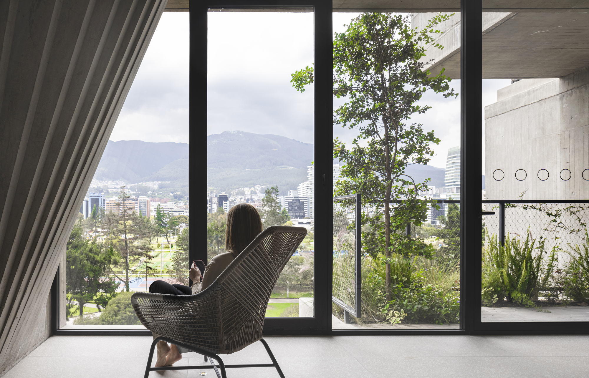 Interior view through floor-to-ceiling windows showing a woven chair facing trees and parkland beyond