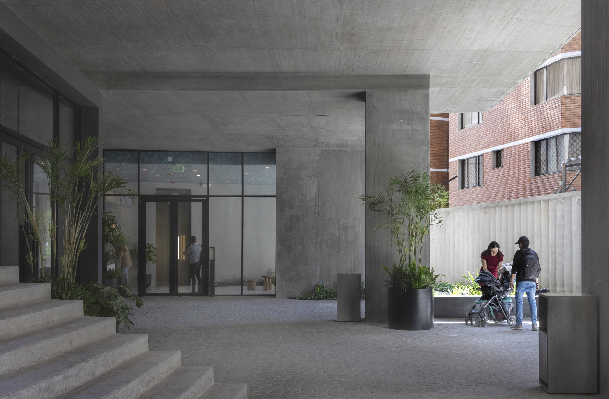 Covered entry courtyard with board-formed concrete ceiling and family with stroller walking past potted palms