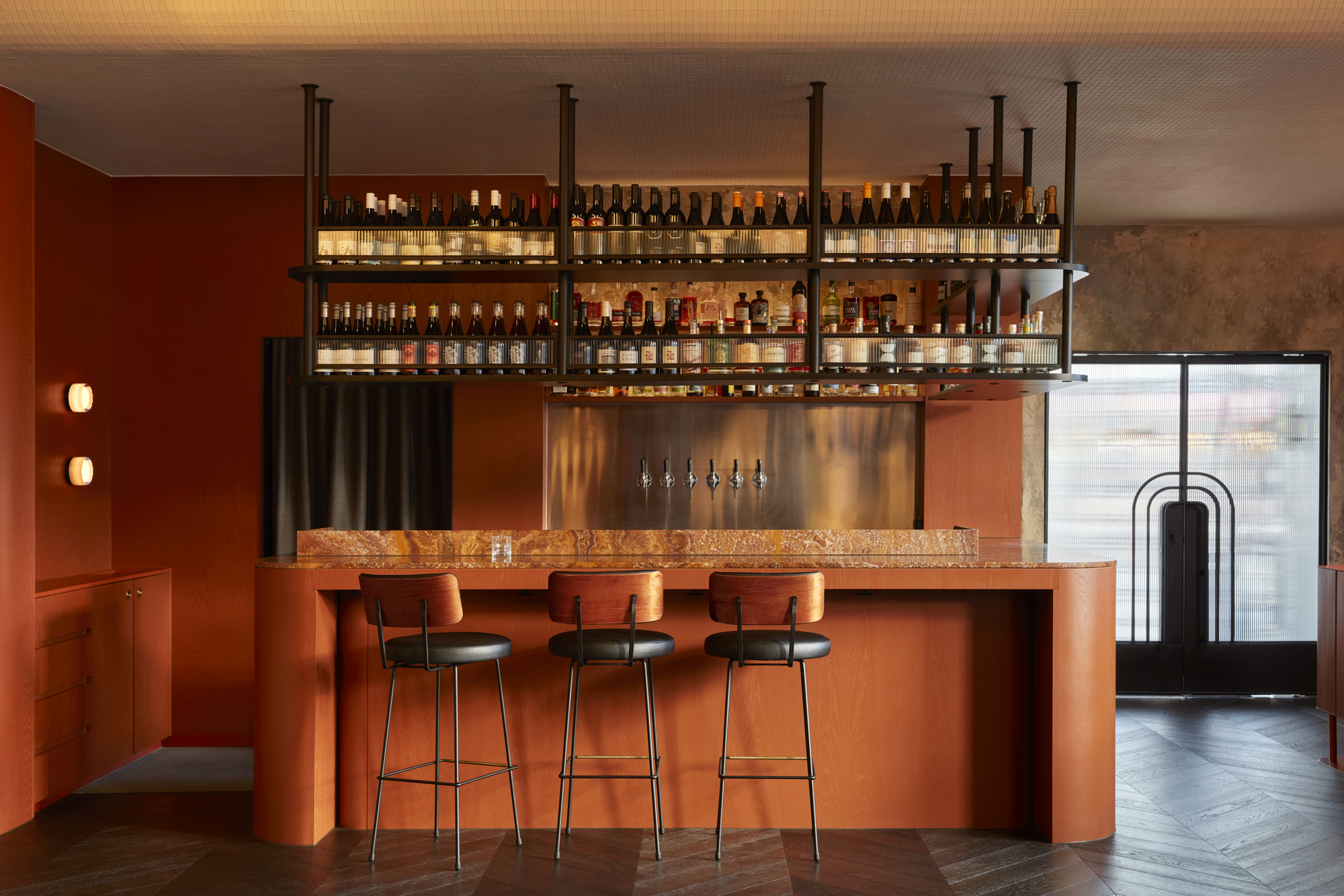 Bar counter with terracotta stone top, suspended shelving, and three bar stools beneath backlit bottles