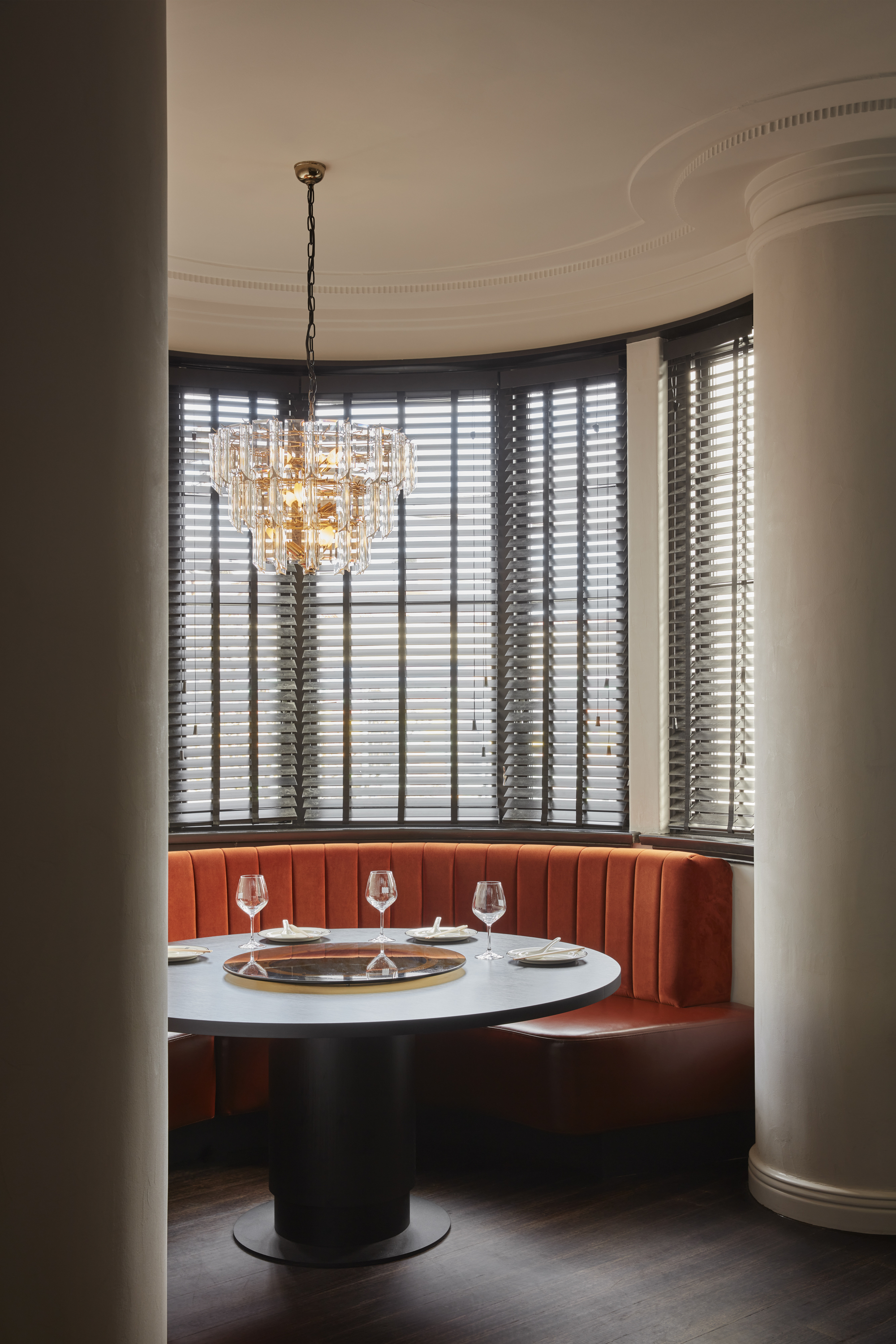 Curved booth with coral banquette, crystal chandelier, and tall windows with venetian blinds at dusk