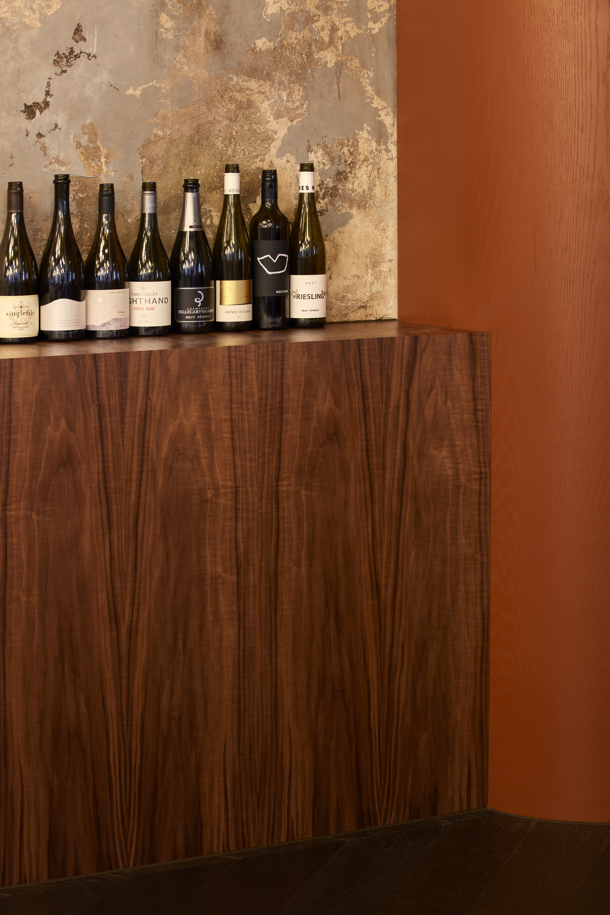 Walnut veneer cabinet with wine bottles displayed against weathered plaster wall in natural light