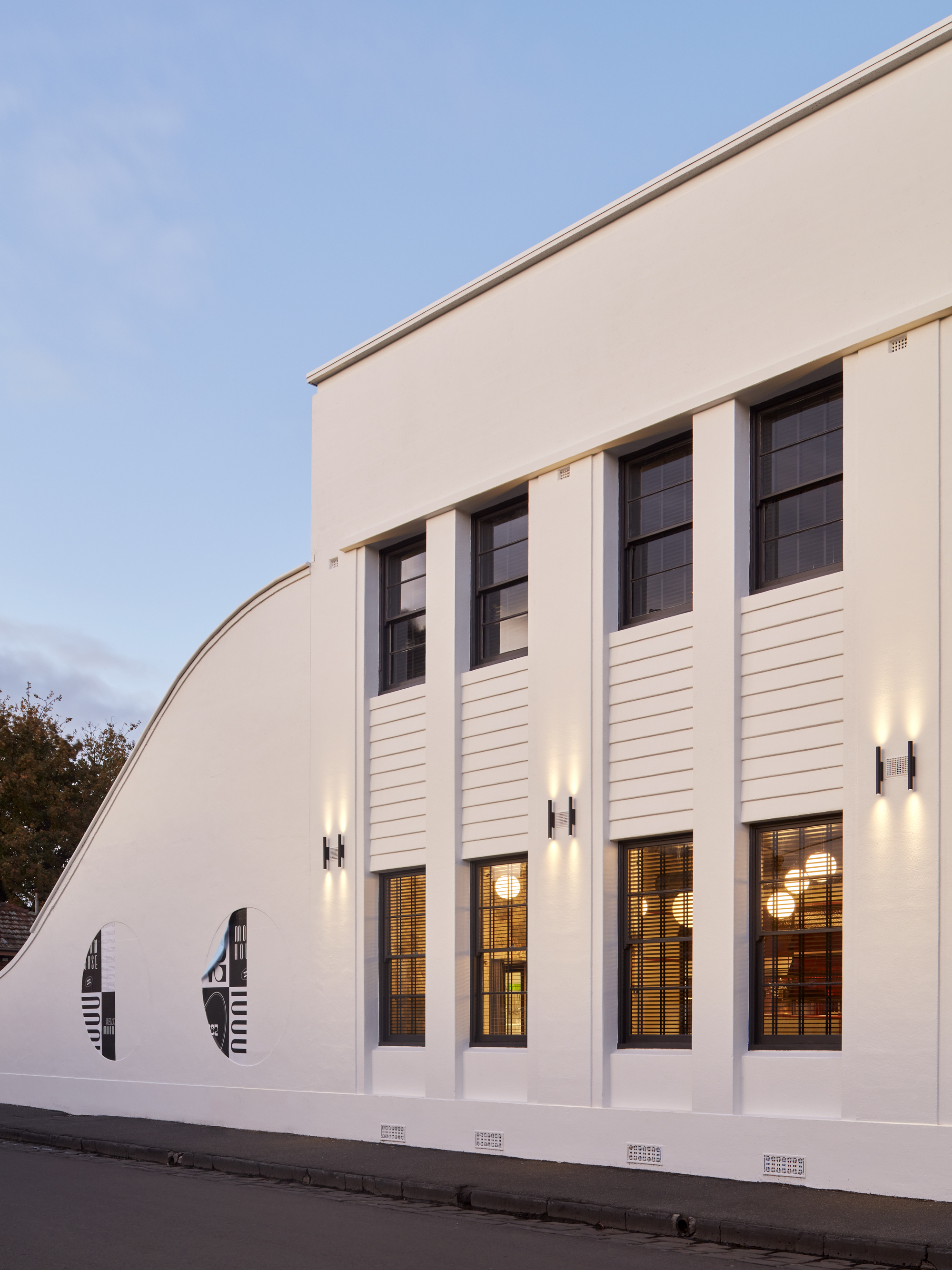White rendered facade with vertical pilasters and clerestory windows illuminated at dusk from the street