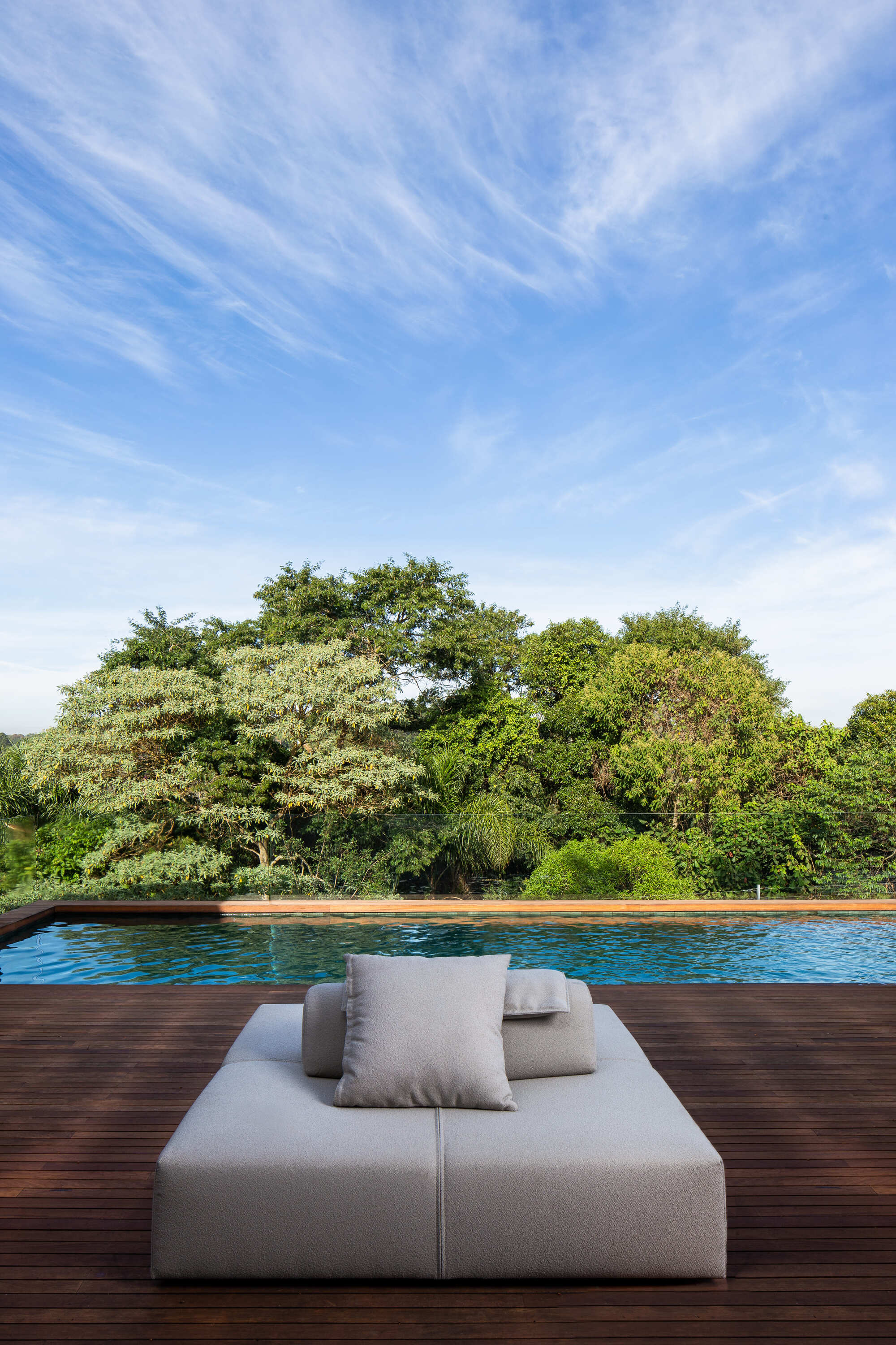 Timber pool deck with cushioned lounger facing mature trees under a blue sky with wispy clouds
