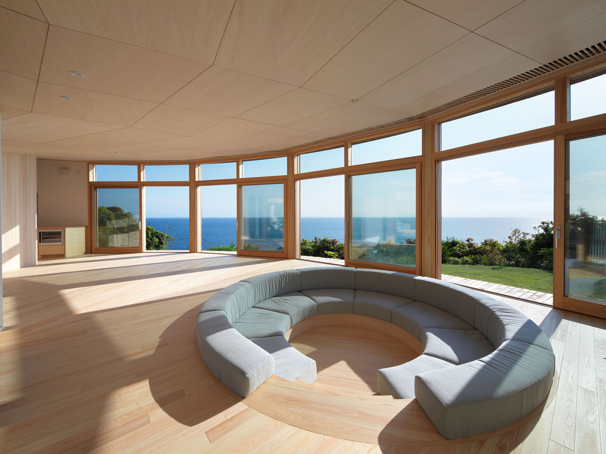 Curved living space with continuous timber-framed windows overlooking the ocean under a plywood ceiling