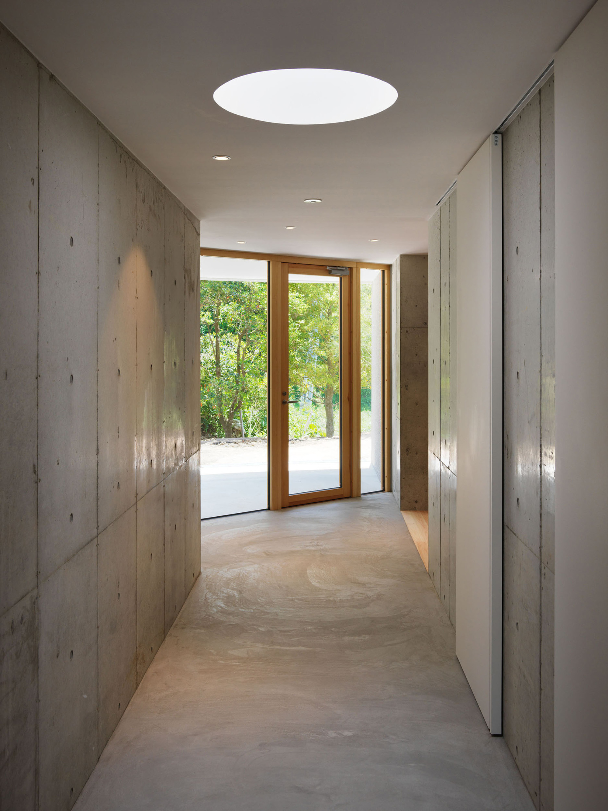 Concrete corridor with circular skylight and timber-framed doors leading to garden