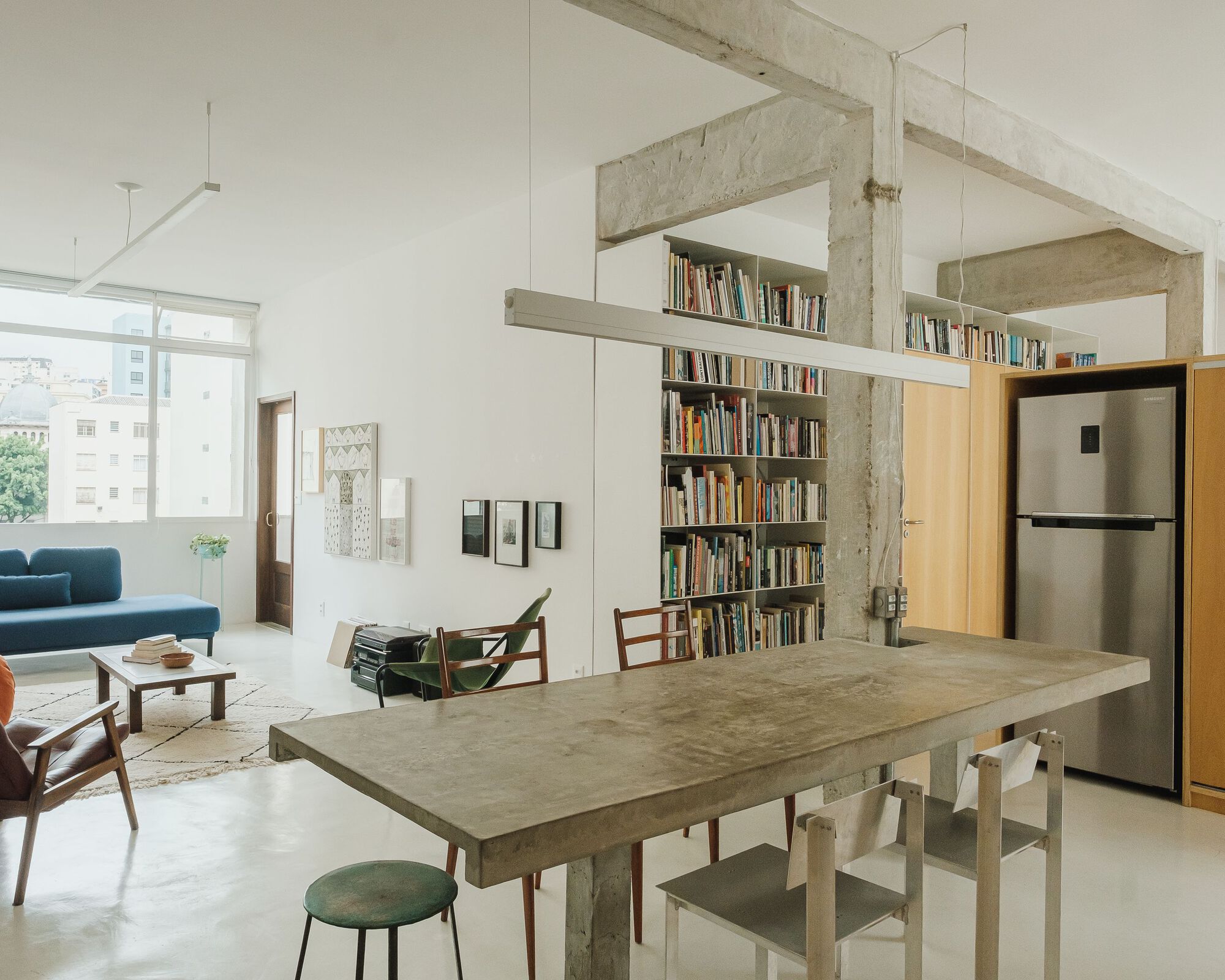 Open living and dining area with exposed concrete frame, bookshelf wall, and light from a large window