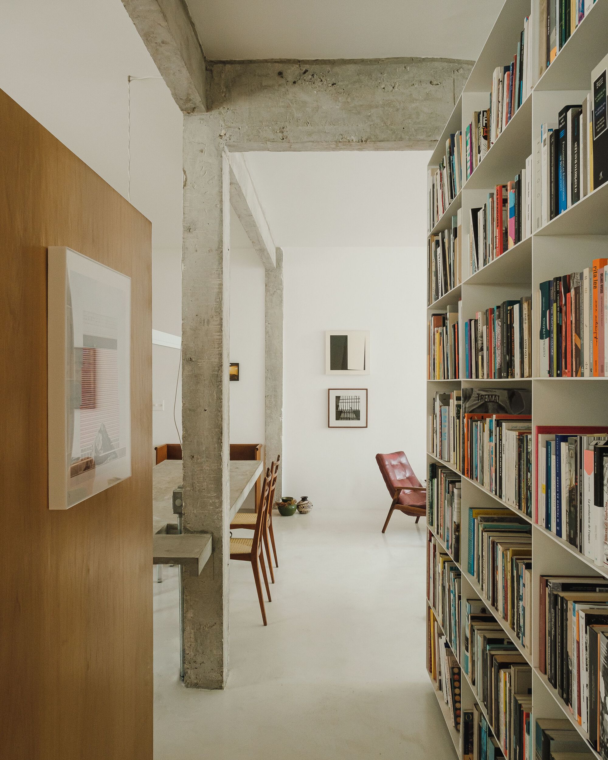 Corridor view past concrete columns toward white bookshelves and a red leather chair