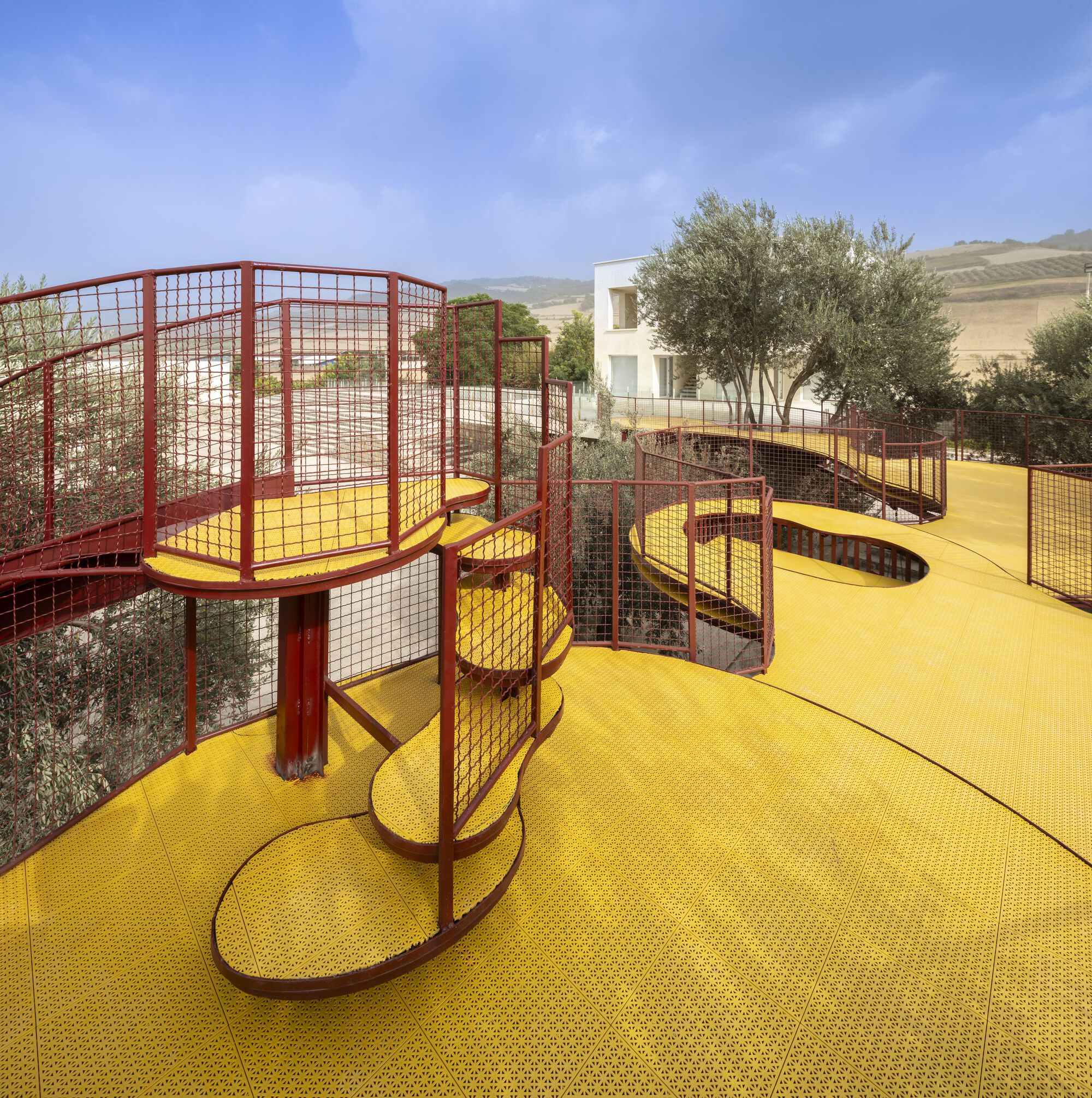 Elevated yellow walkway with red mesh railings winding between olive trees under a clear blue sky