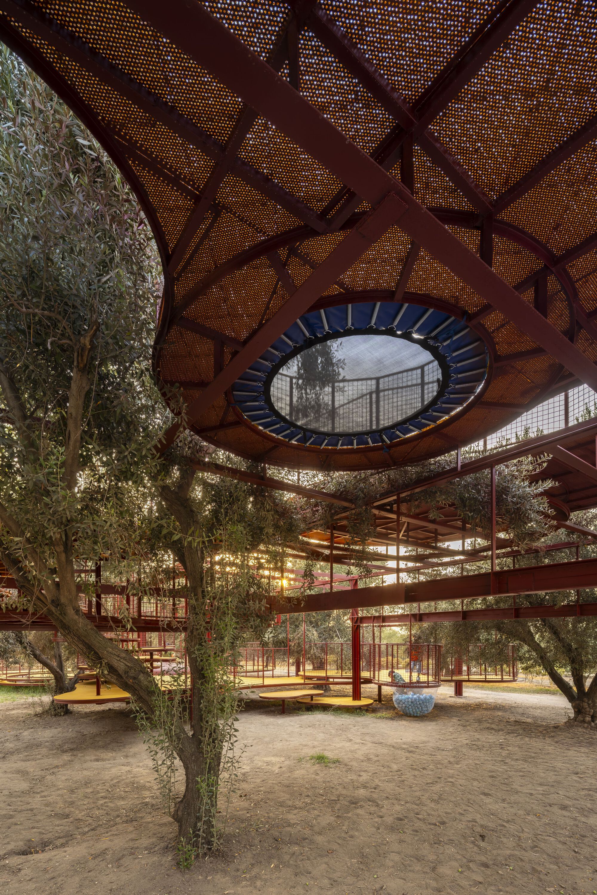 Underside view of circular shade canopy with red perforated metal mesh and central oculus above olive grove
