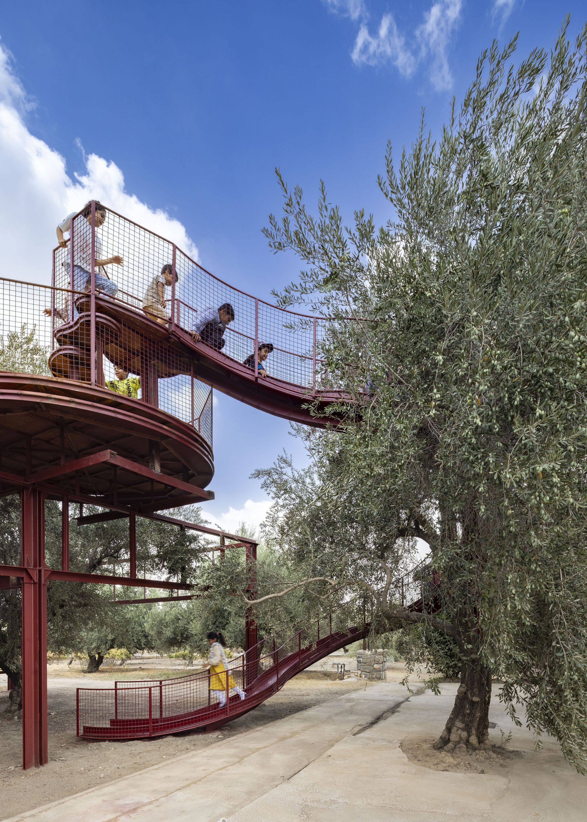 Red steel bridge with children crossing overhead connecting platform levels among olive trees on sunny day