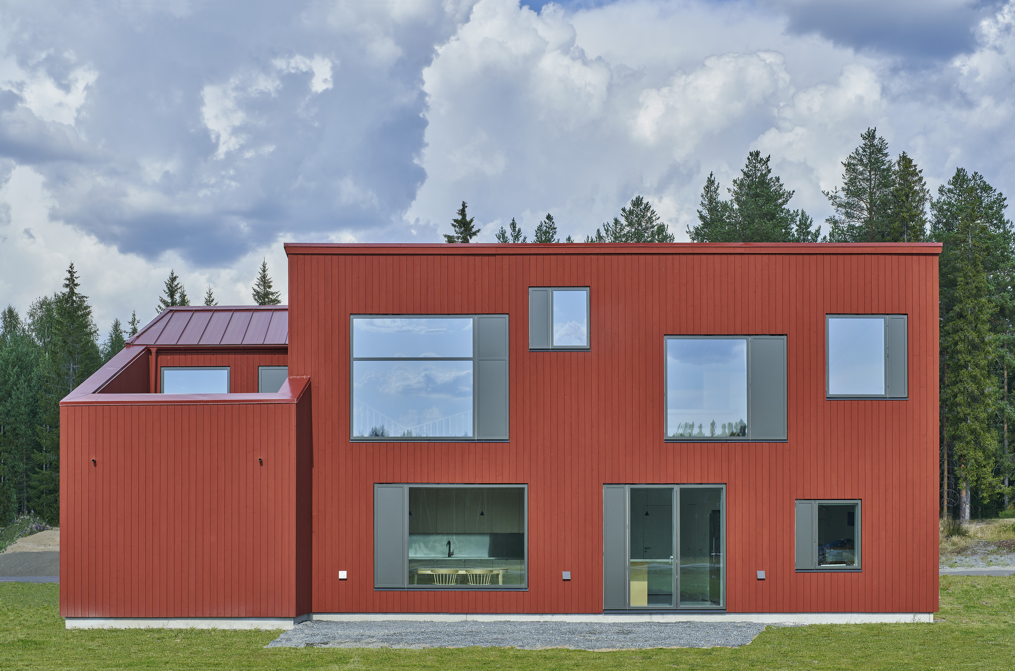 Facade with asymmetrically placed windows in the red vertical cladding overlooking a gravel yard