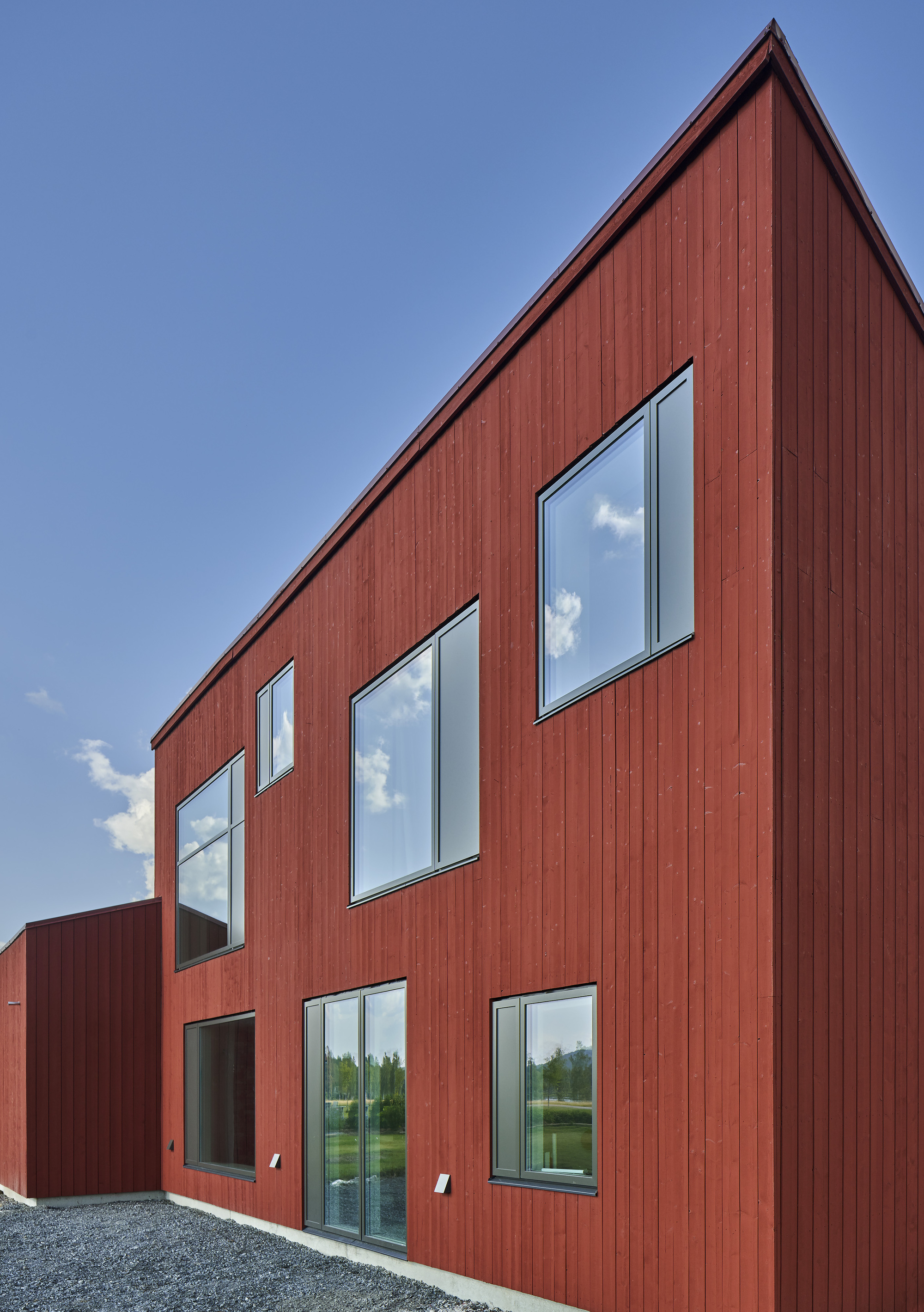 Red vertical wood siding facade with six punched windows reflecting clouds against a clear blue sky