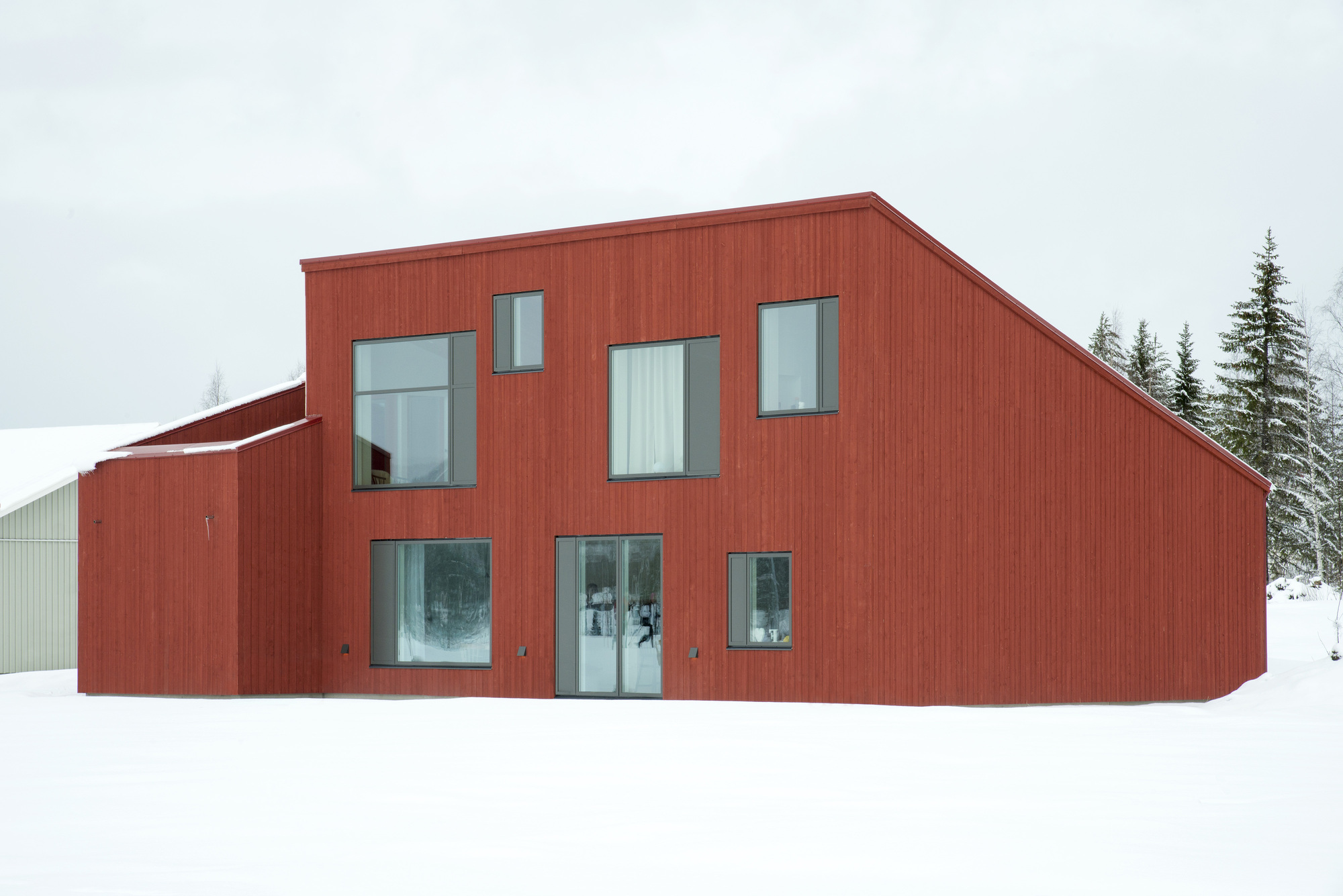 Two-story red cladded volume with asymmetrical windows sitting in deep snow beside a distant structure
