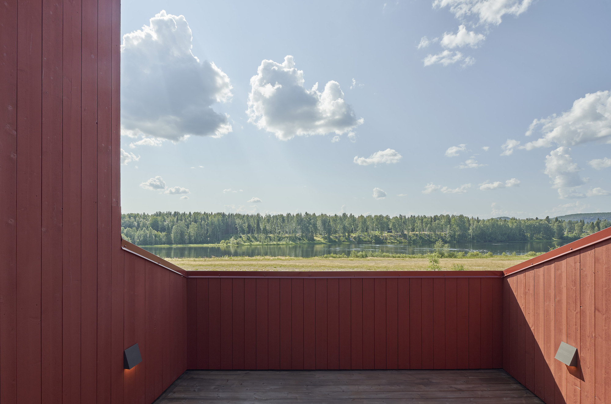 Roof terrace enclosed by red wood paneling framing a lake and forest under a cloudy summer sky