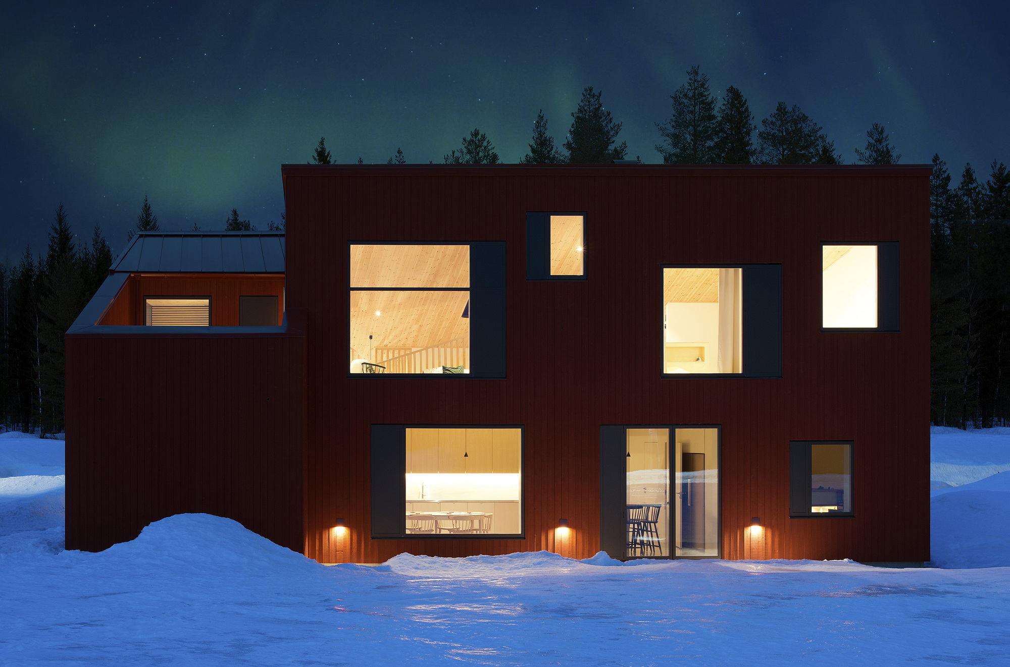 Illuminated windows at dusk reveal interior spaces while three wall-mounted lights glow against snowbanks