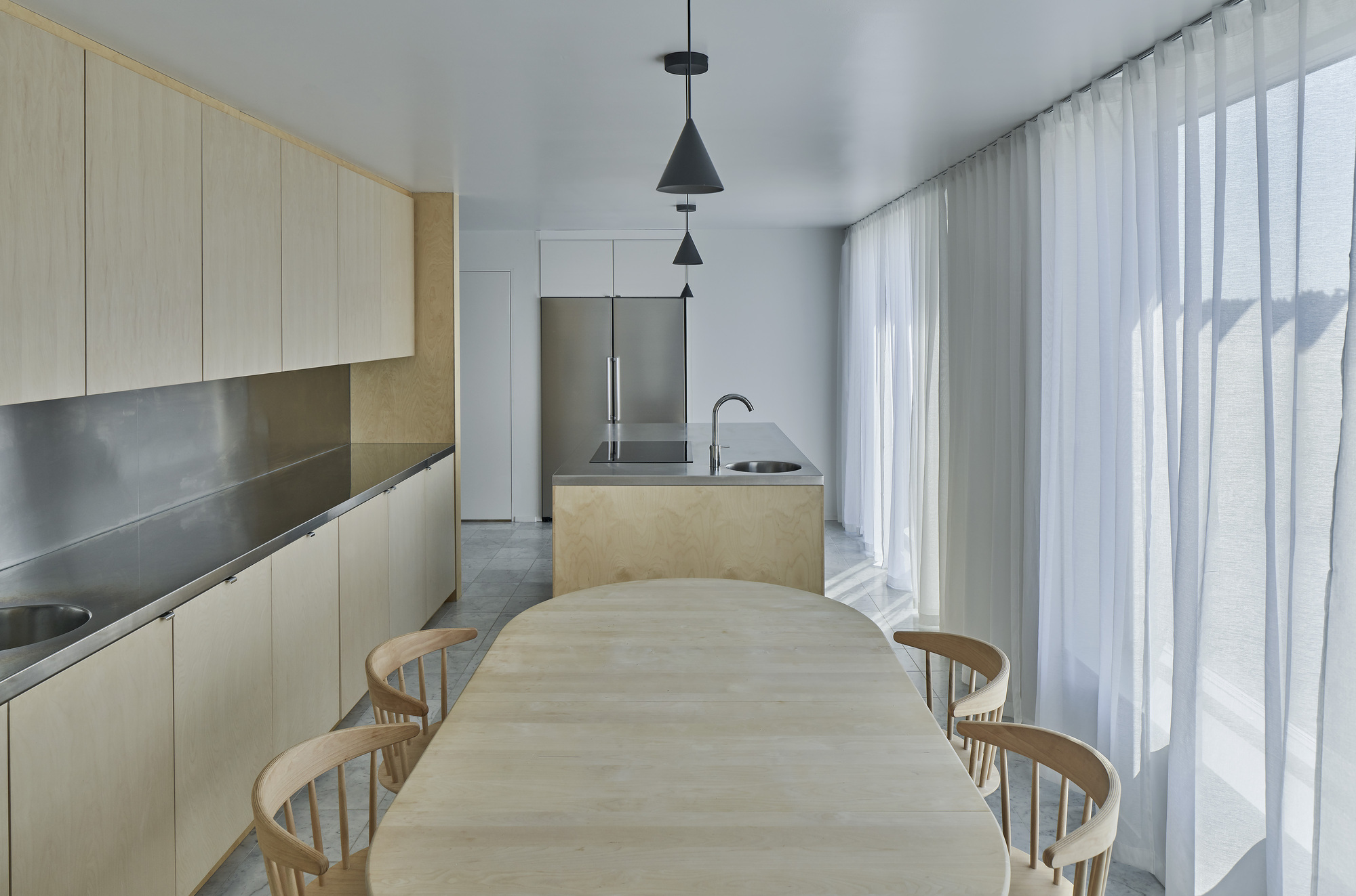 Open kitchen and dining area with pale timber cabinetry, stainless steel countertops, and sheer curtains filtering daylight