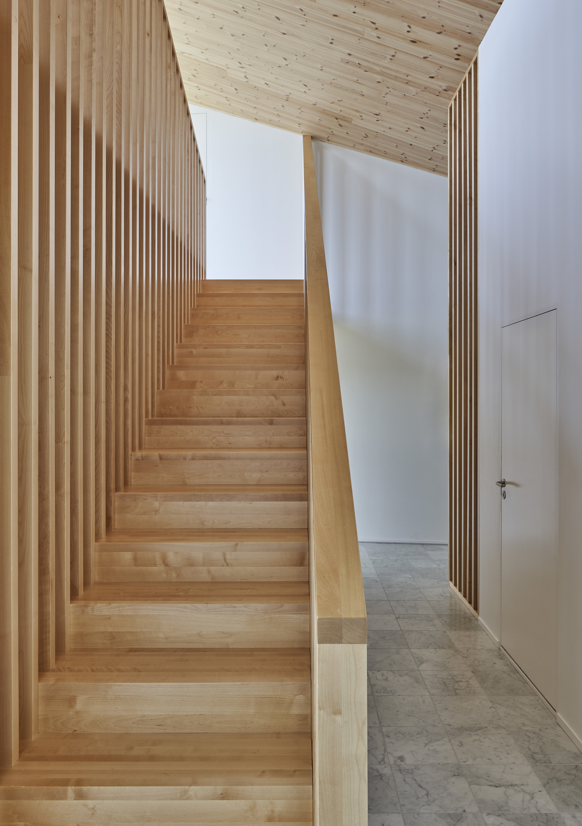 Timber staircase with vertical slat balustrade ascending beneath an angled plywood ceiling