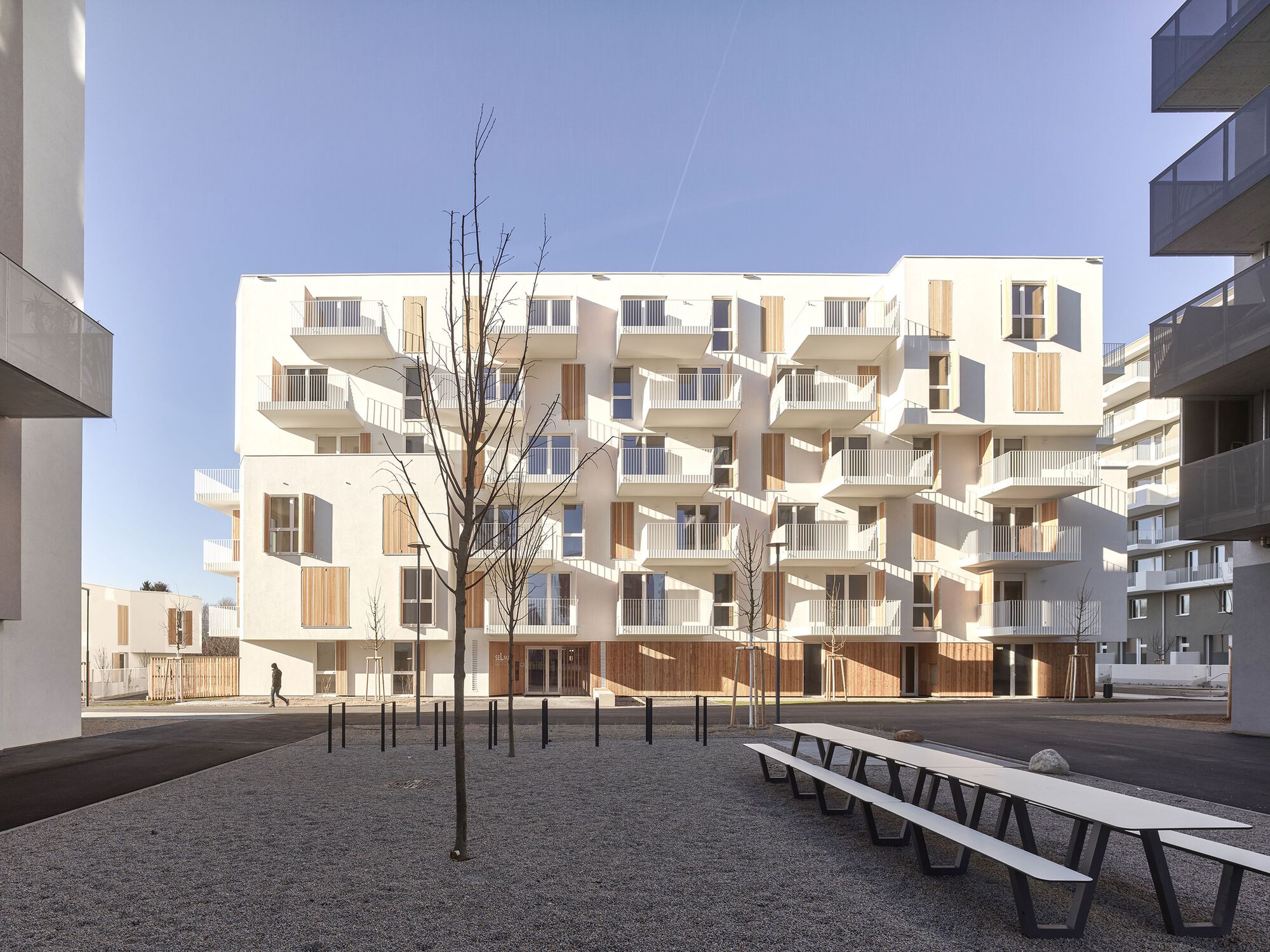 Street facade with projecting white balconies and timber screens beside bare winter trees