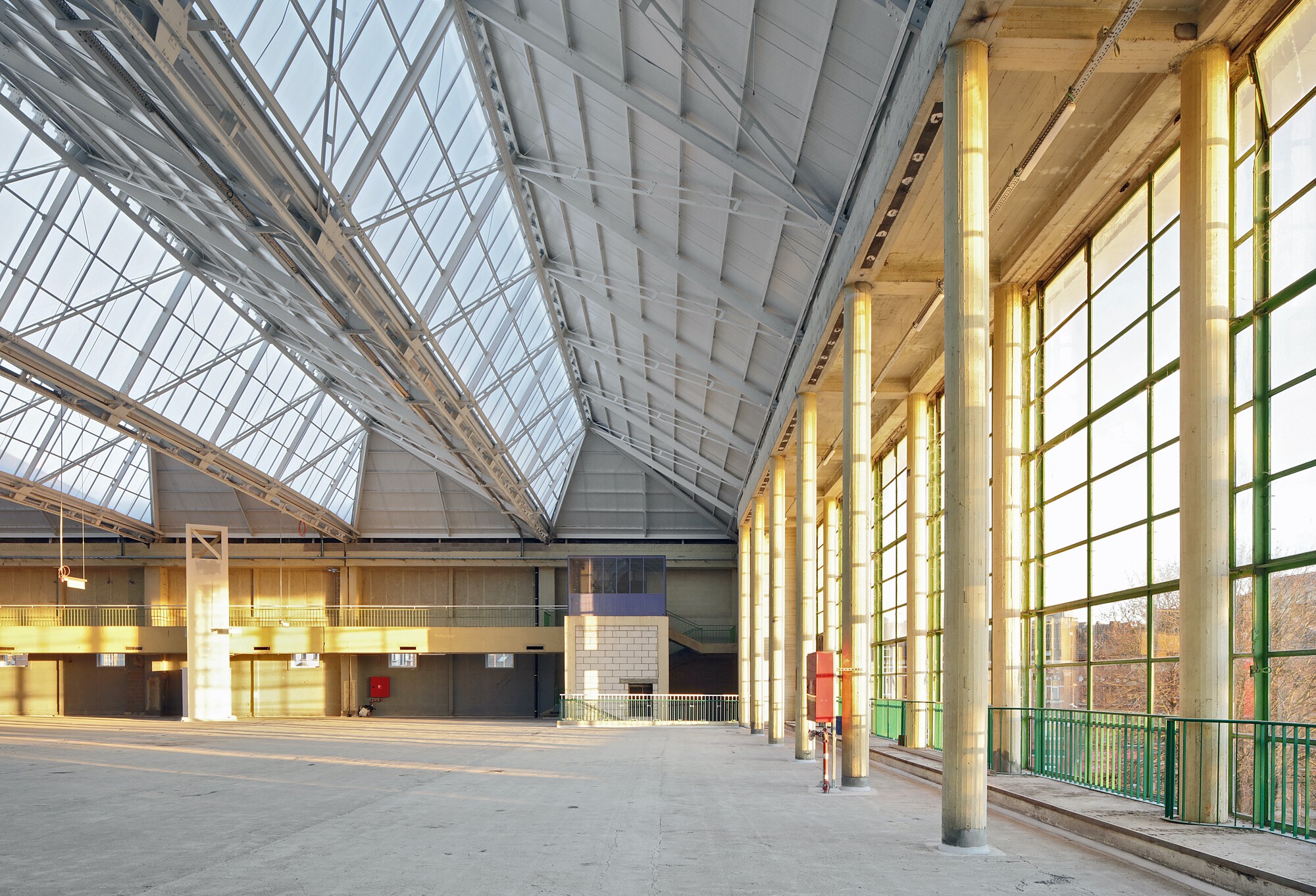 Interior hall with angled steel roof glazing and timber columns along gridded windows in afternoon light