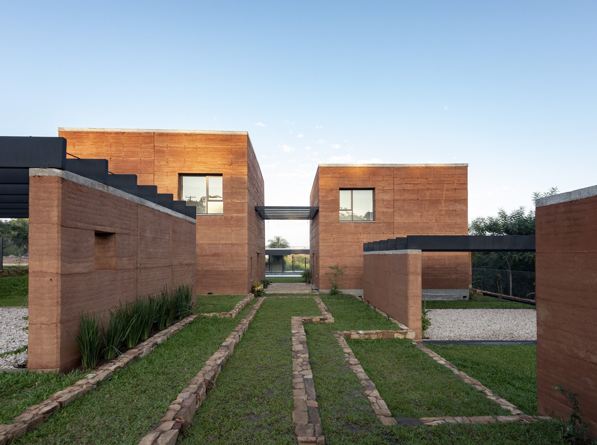 Front courtyard with rammed earth walls, planted grass strips and gravel paths leading to a skybridge