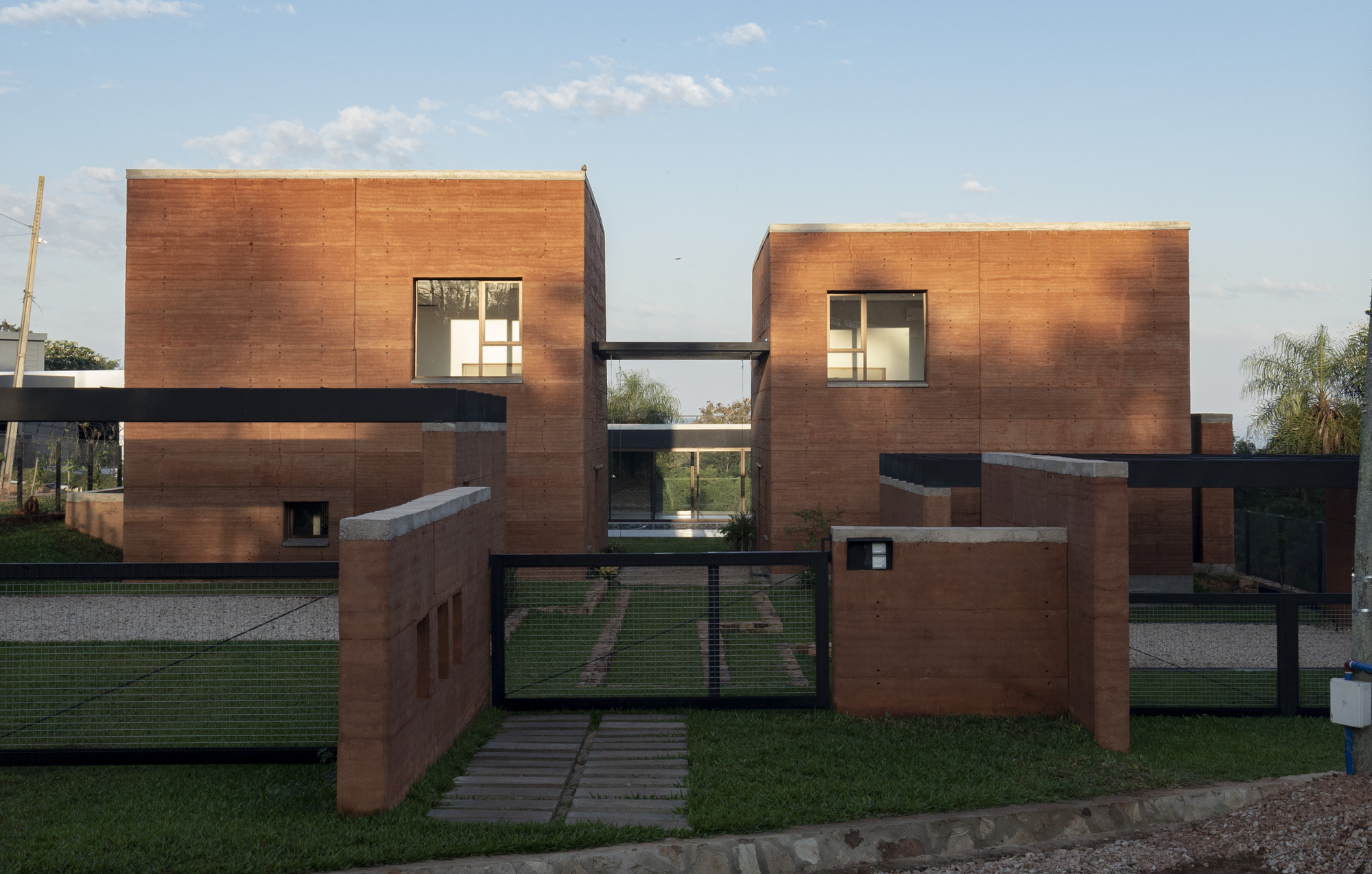 Street view of paired rammed earth volumes connected by a bridge under evening light